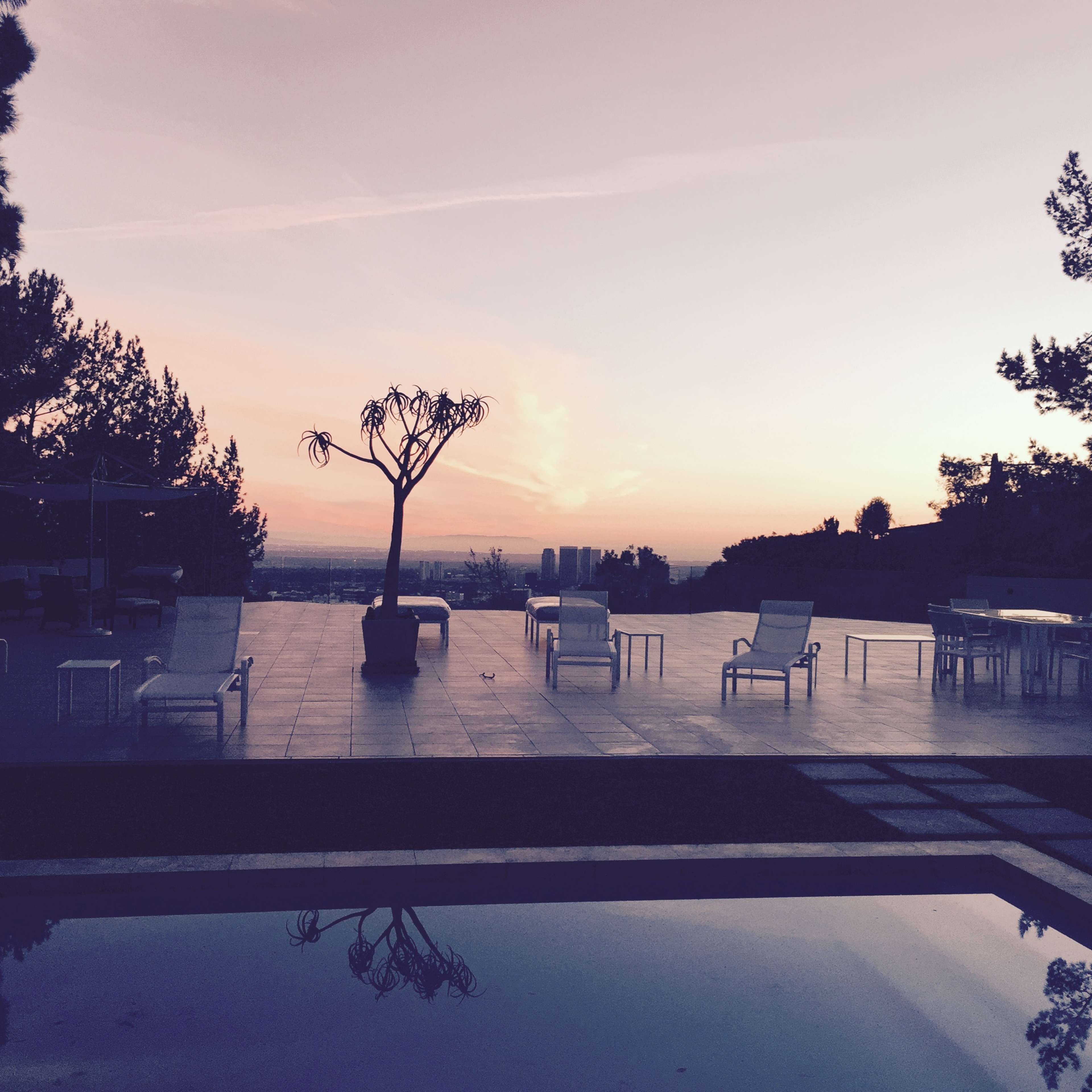 A patio area with white lounge chairs and tables, overlooking a sunset with a pool reflecting the scene.