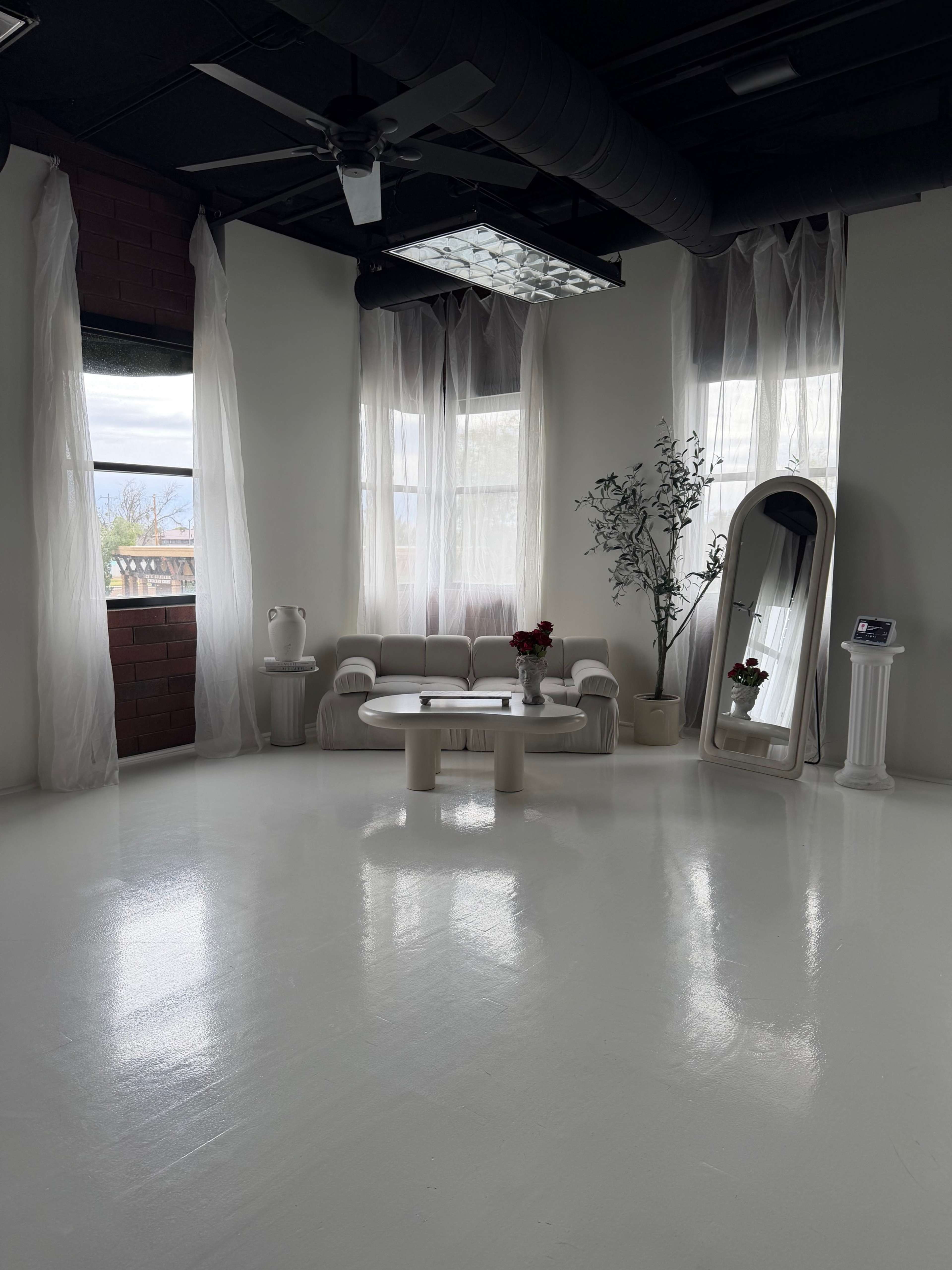 The image shows a modern, minimalistic interior space with large windows, sheer curtains, a circular coffee table, a mirror, and a potted plant.