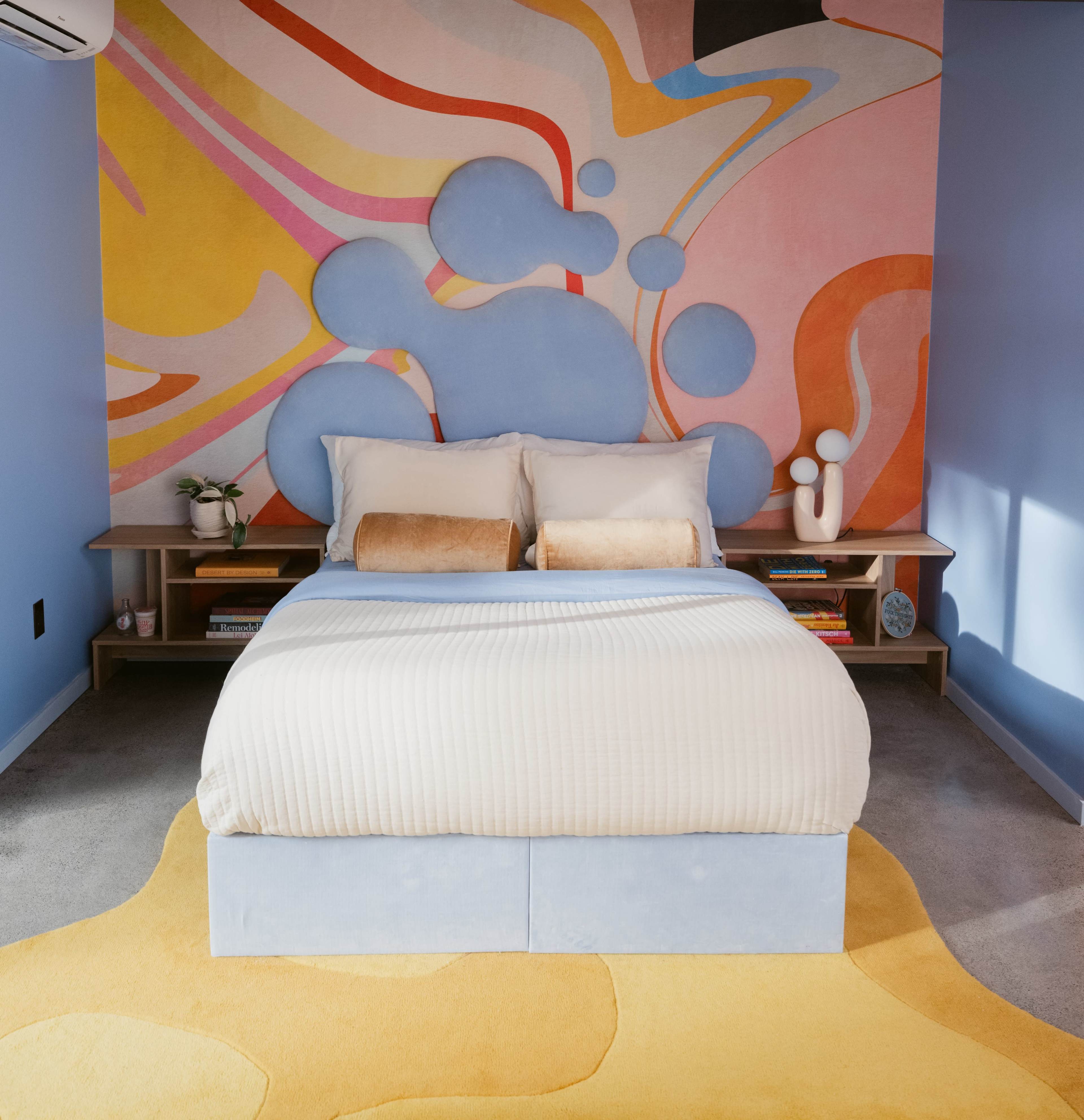 The image shows a modern bedroom with a bed centered against a colorful abstract mural and light-colored carpet.