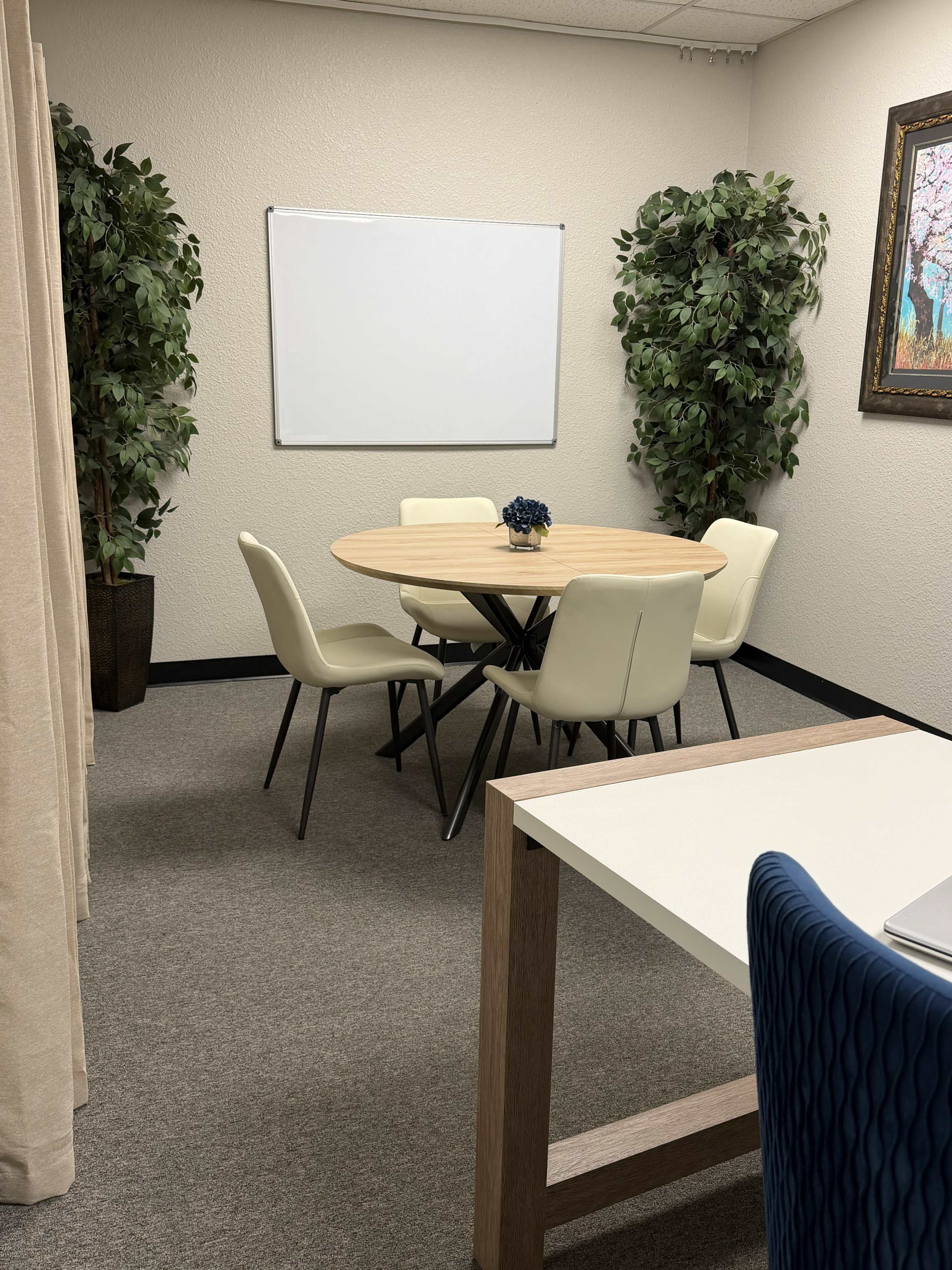 The image shows a small meeting room with a round wooden table surrounded by five white chairs, flanked by two tall green plants and a blank whiteboard on the wall.