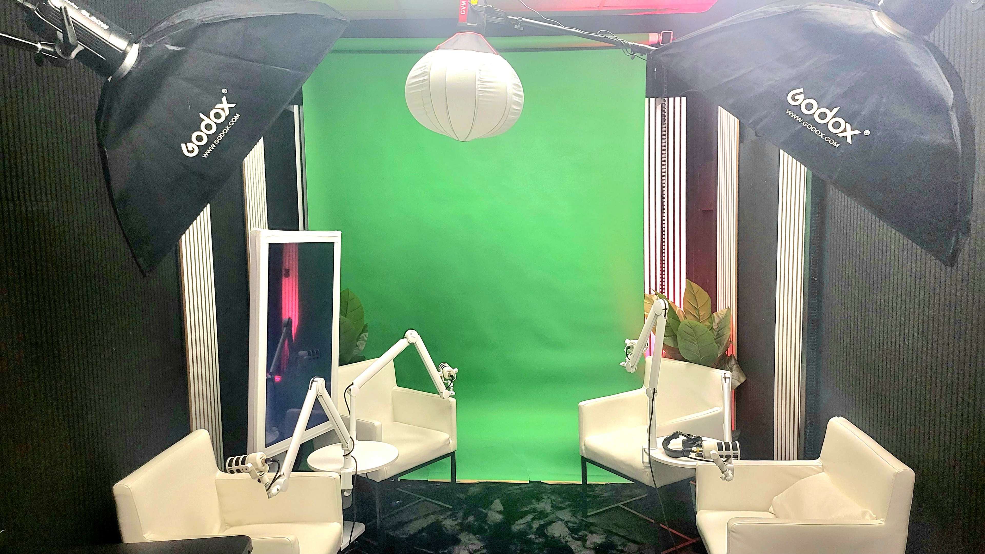 The image shows a small studio setup with two white chairs, a round table, and a green screen, surrounded by lighting equipment.