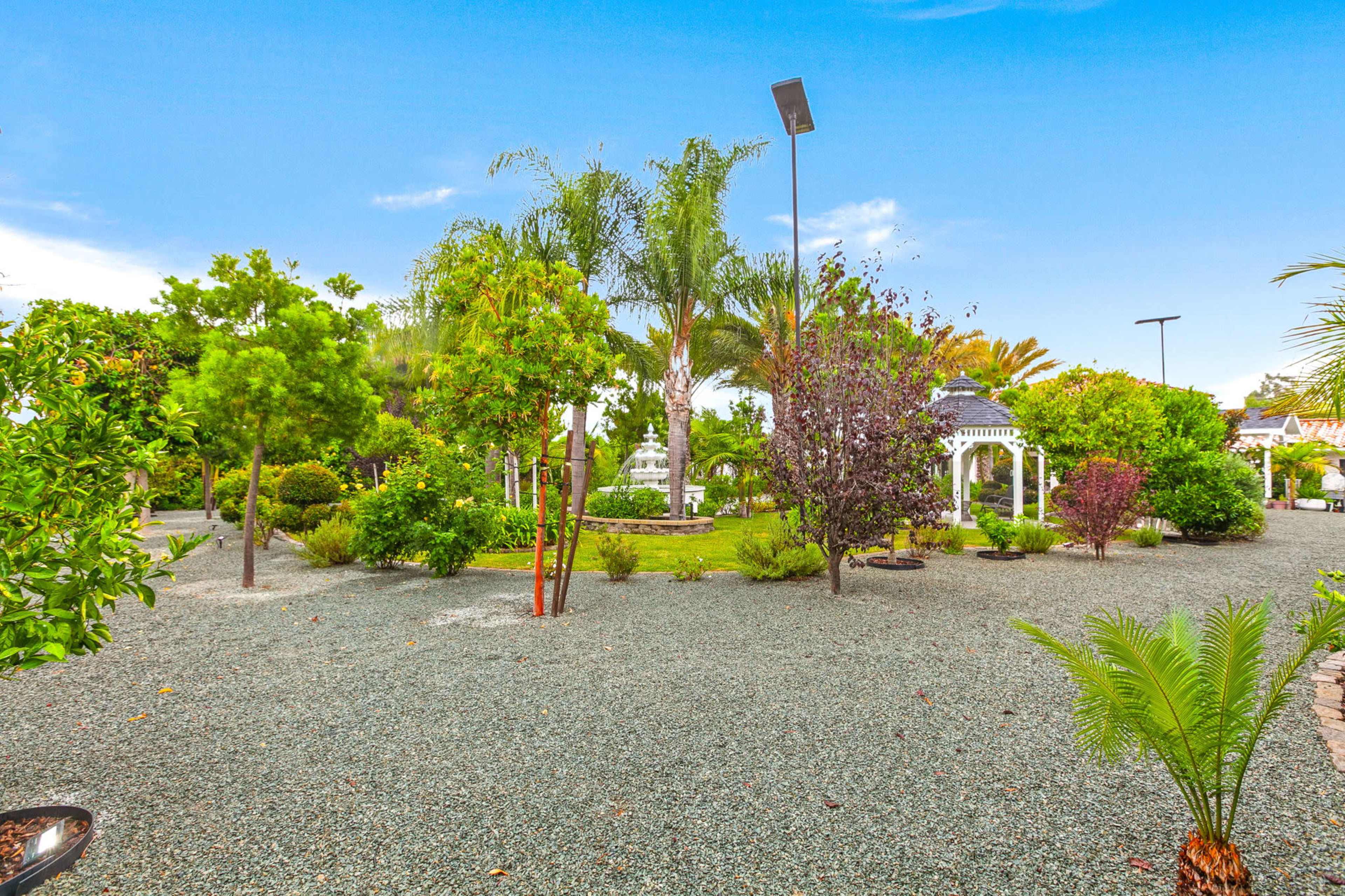 The image shows a landscaped garden featuring various trees and shrubs, a gravel path, and a decorative gazebo.
