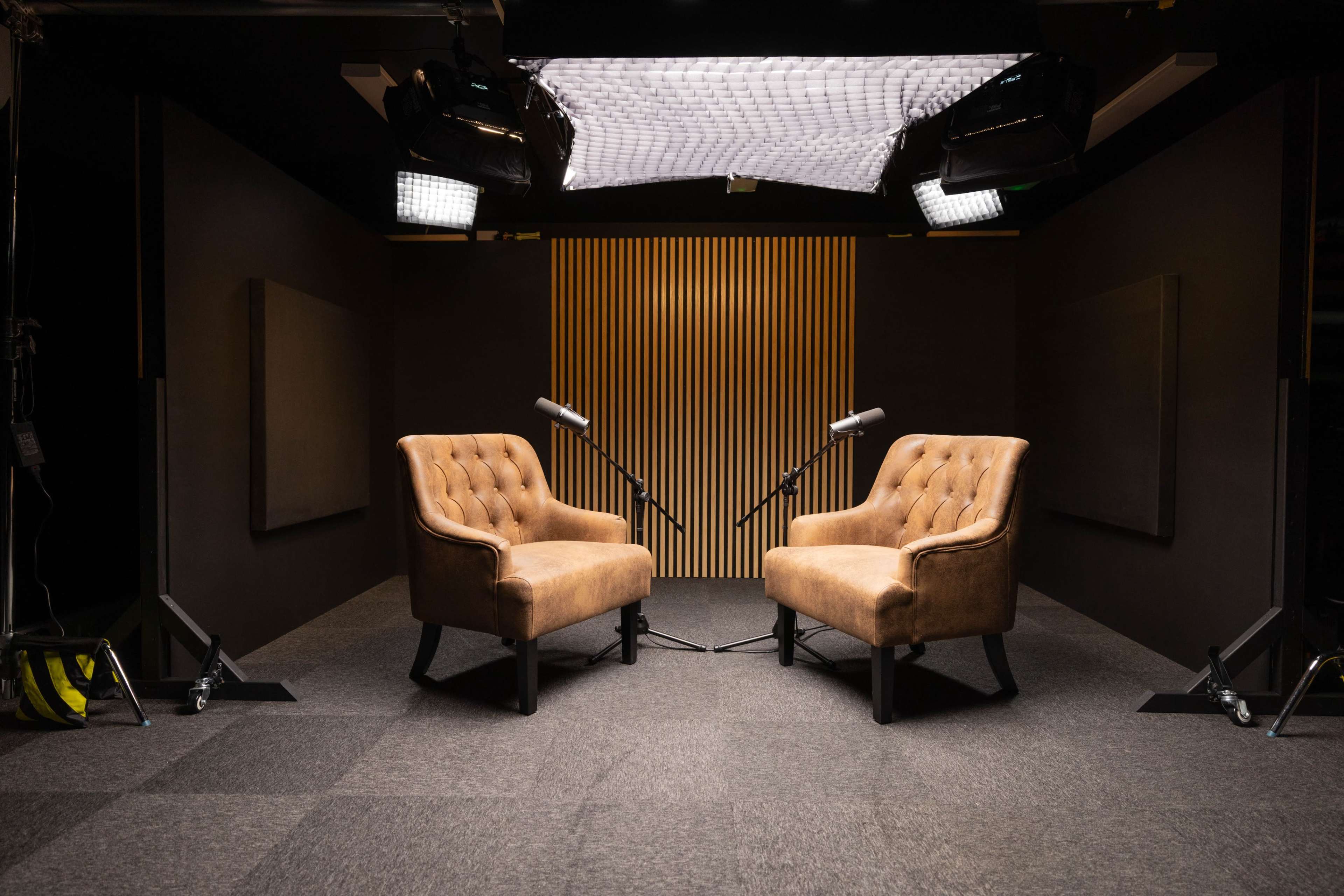 The image shows two upholstered chairs facing each other in a soundproof studio, with microphones positioned between them and lighting equipment overhead.