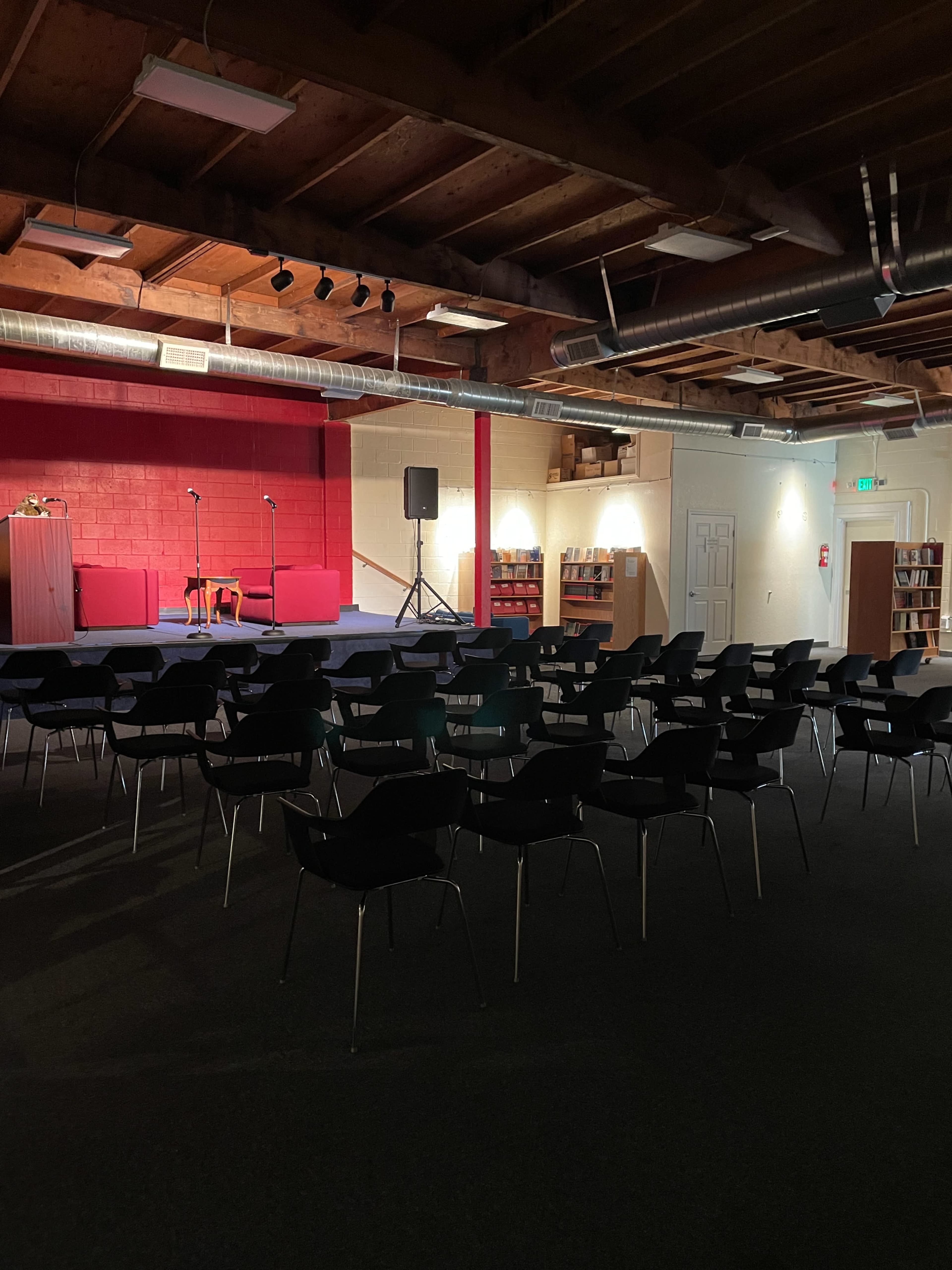A dimly lit room with a small stage, two microphones on stands, and rows of black chairs facing the stage.