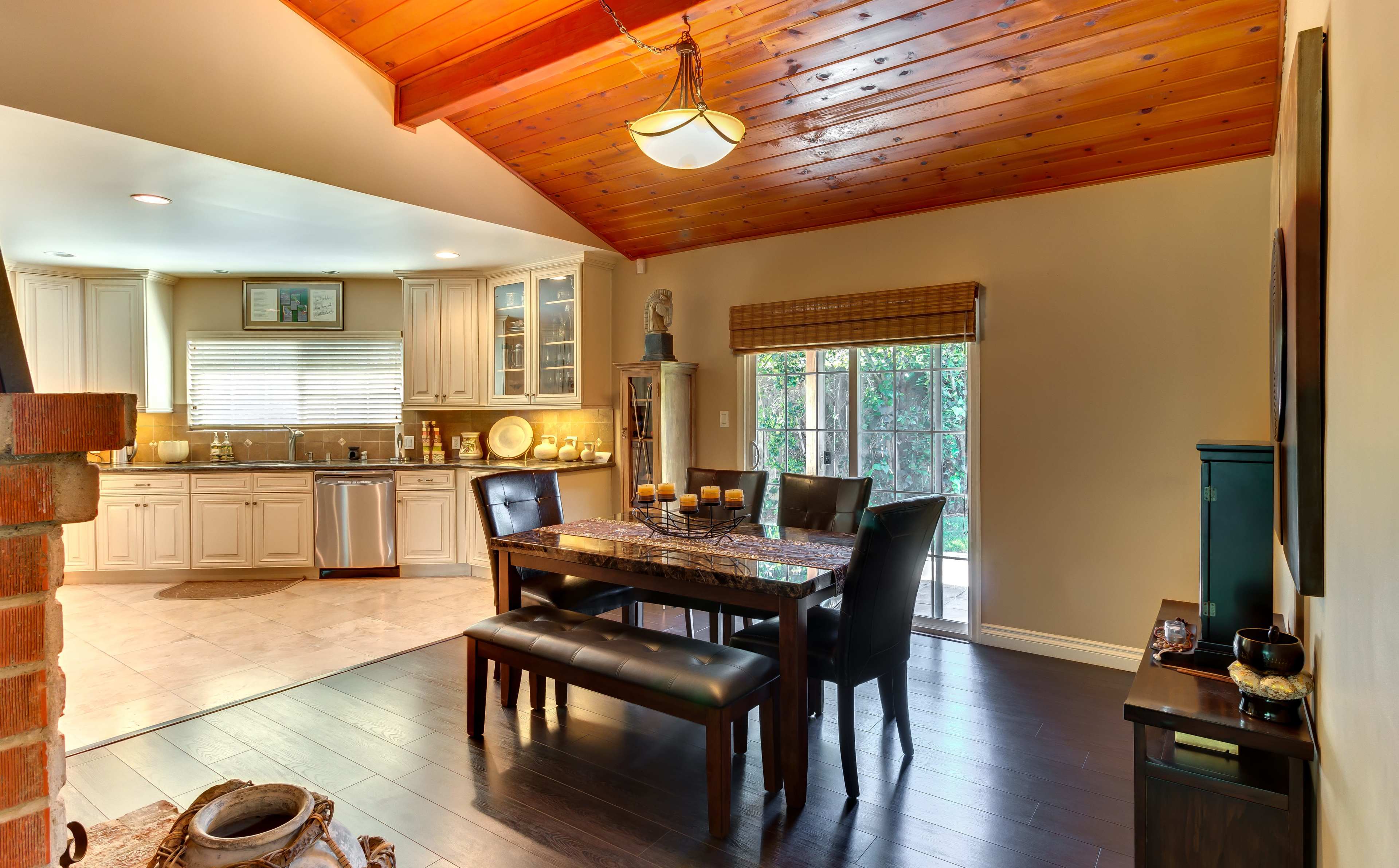 The image shows a dining area with a wooden table and black leather chairs, adjacent to a kitchen with white cabinets and a large window.