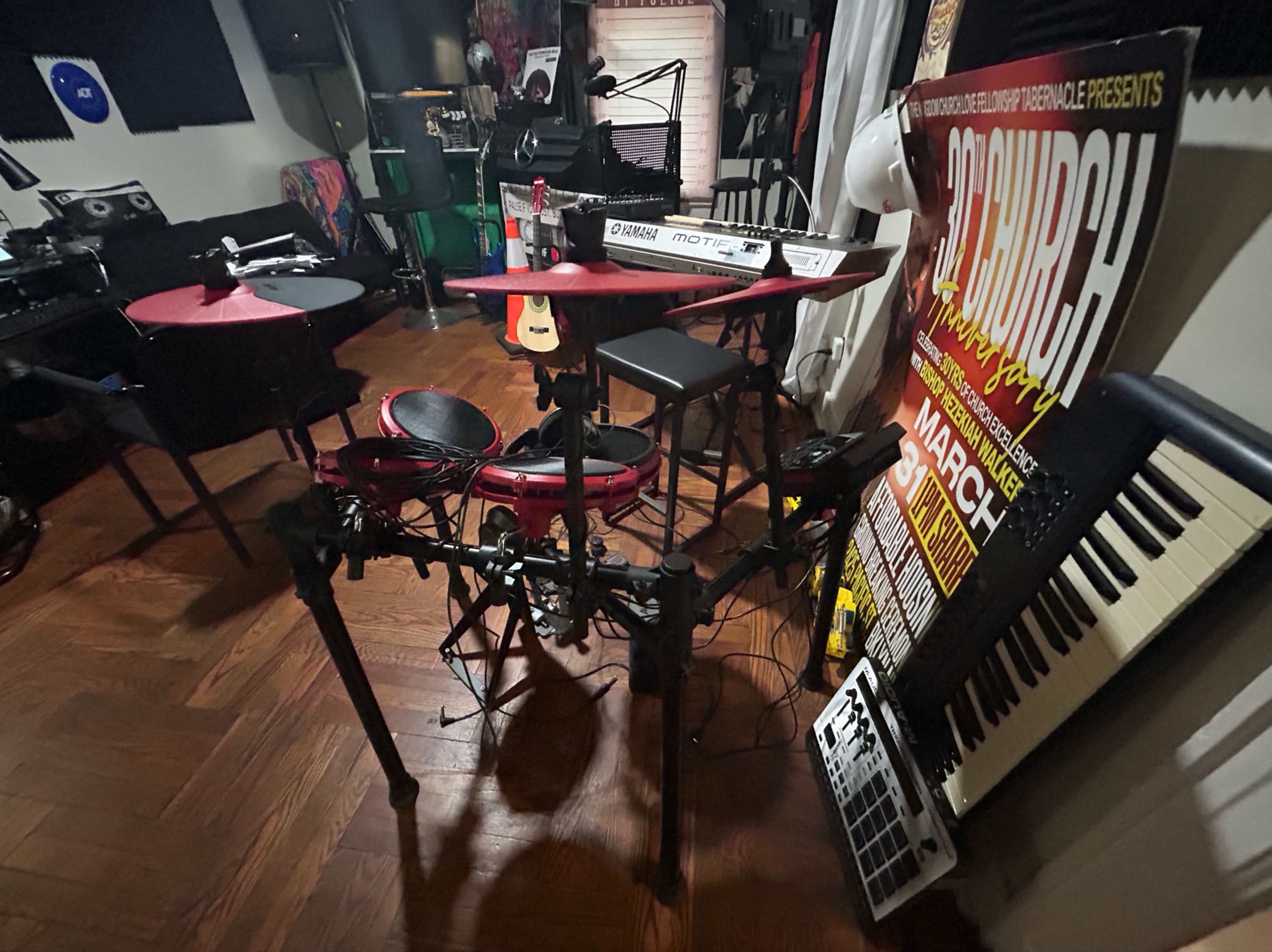 The image shows a music studio with electronic drums, keyboards, and various instruments, along with a promotional poster on the wall.
