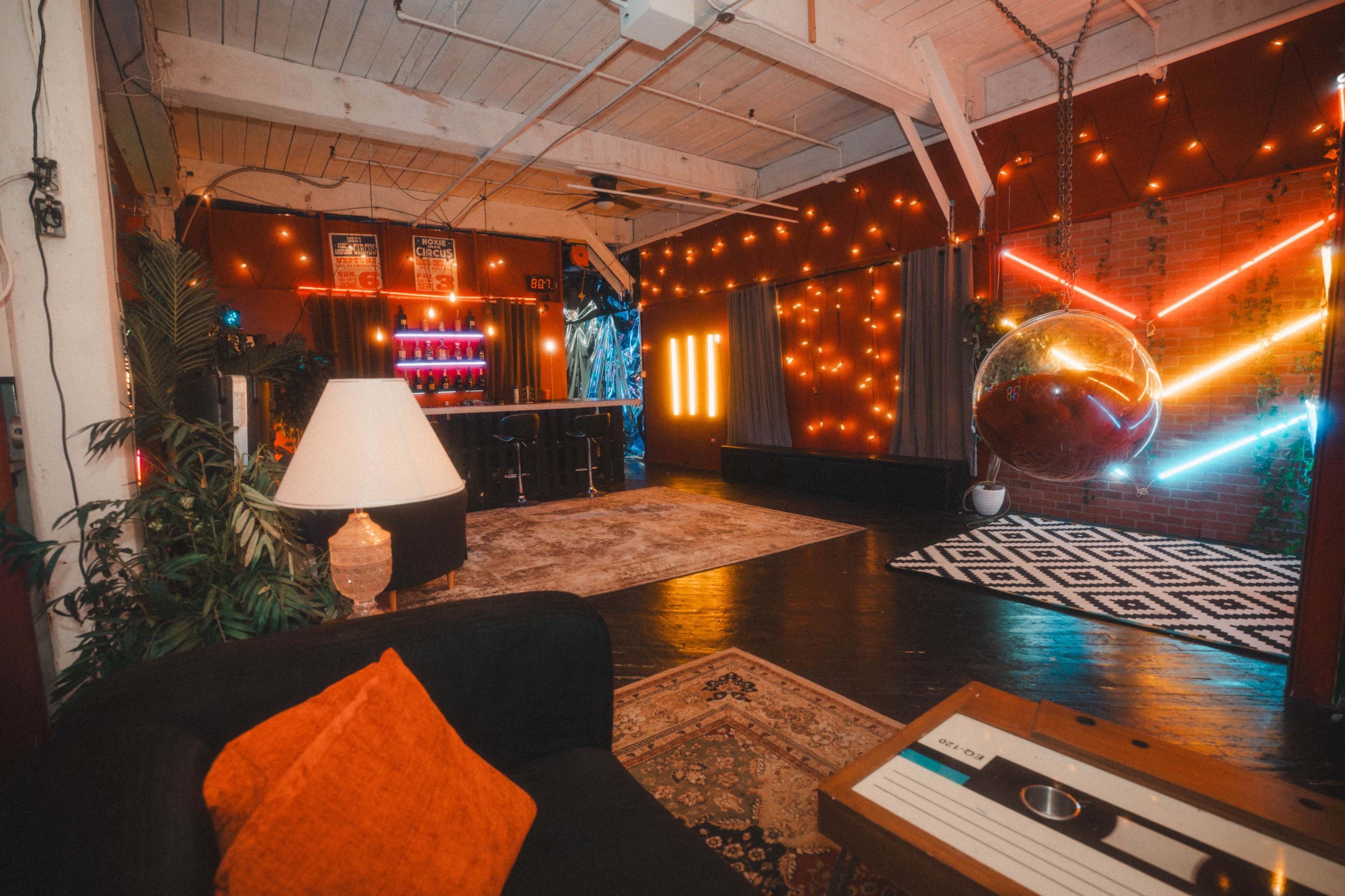 The image shows a spacious indoor lounge area decorated with colorful string lights, a hanging chair, and a patterned rug.