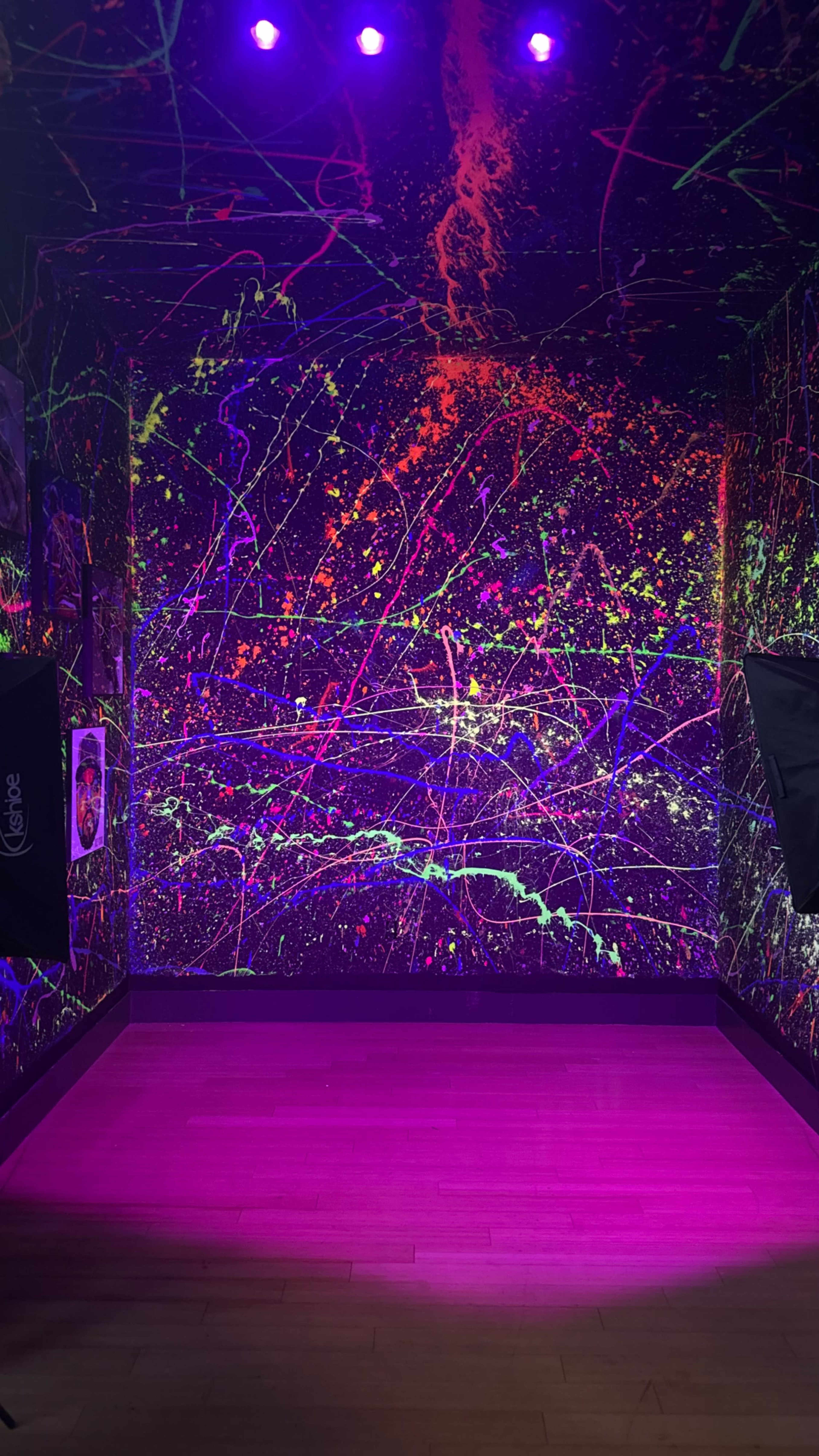 The image shows an indoor space with walls covered in bright, abstract paint splashes and a wooden floor illuminated by purple lighting.