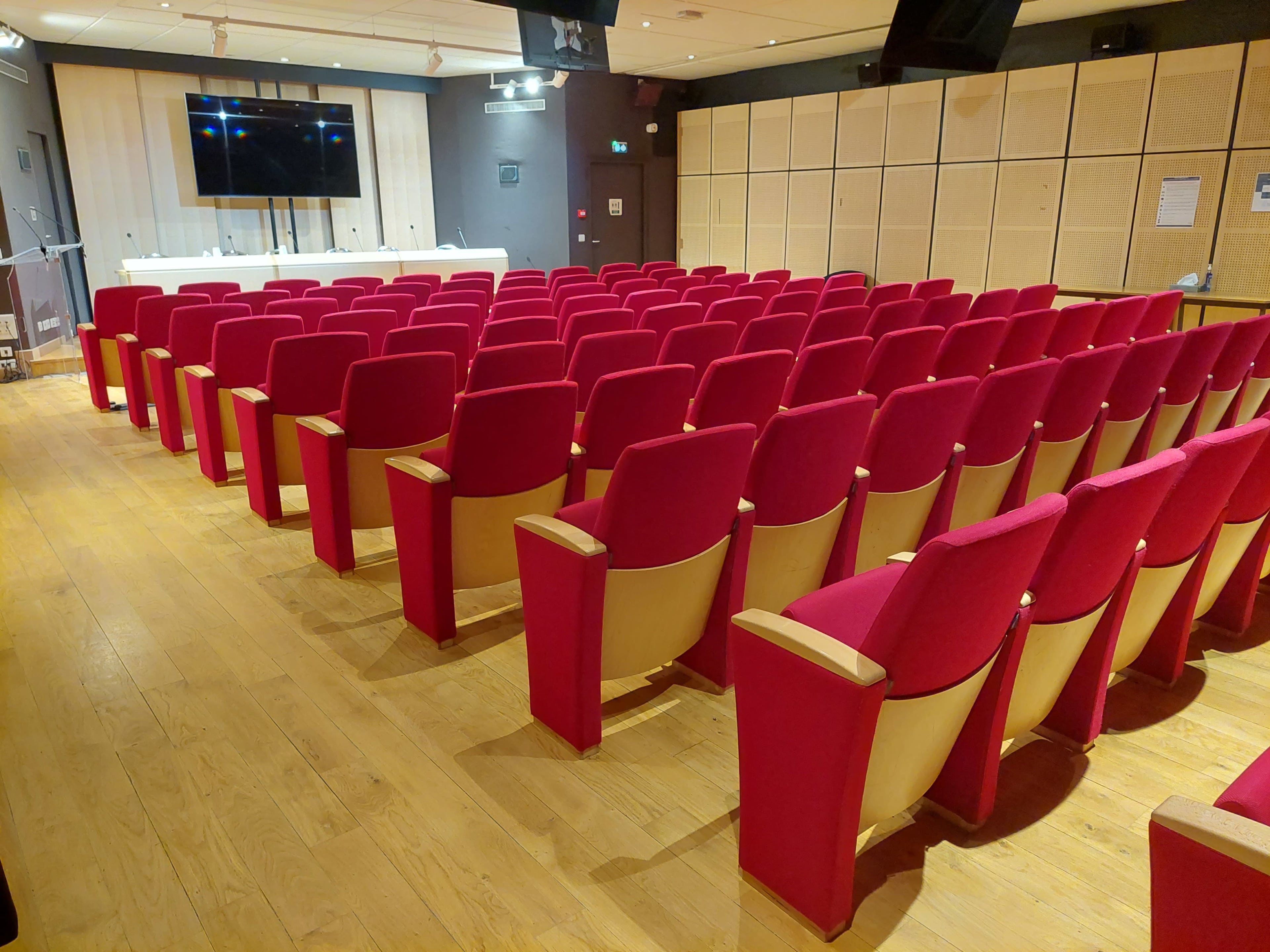 A red upholstered seating area is arranged in rows facing a blank screen in a conference room.