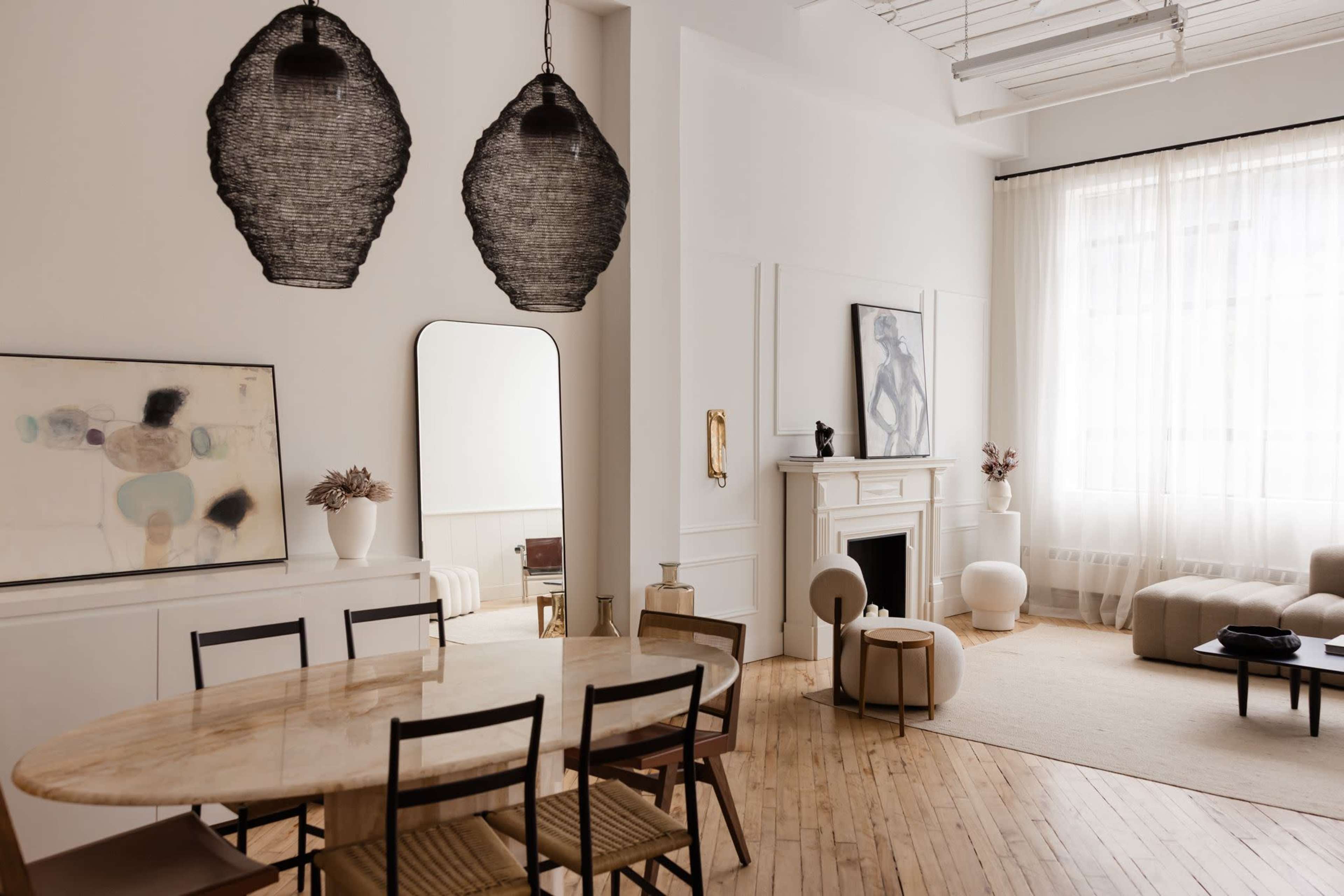 The image shows a modern, minimalist living space with a round dining table, black chairs, a large mirror, and soft furnishings, illuminated by natural light from large windows.