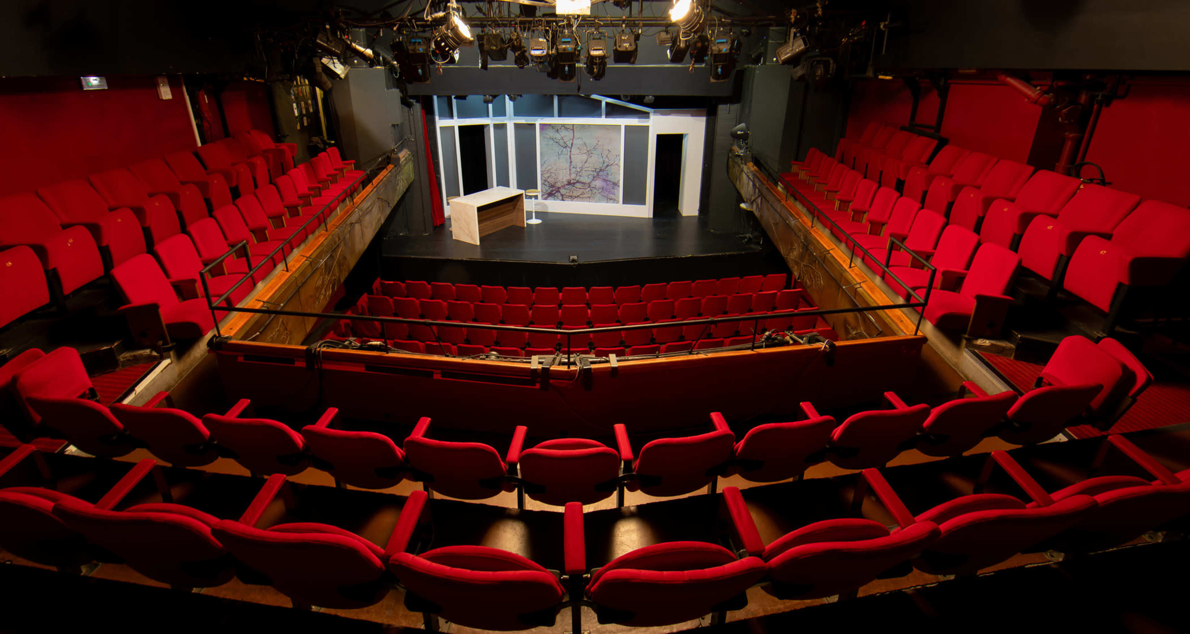 An empty theater interior features red plush seats arranged in a semicircle around a small stage with a simple set design.