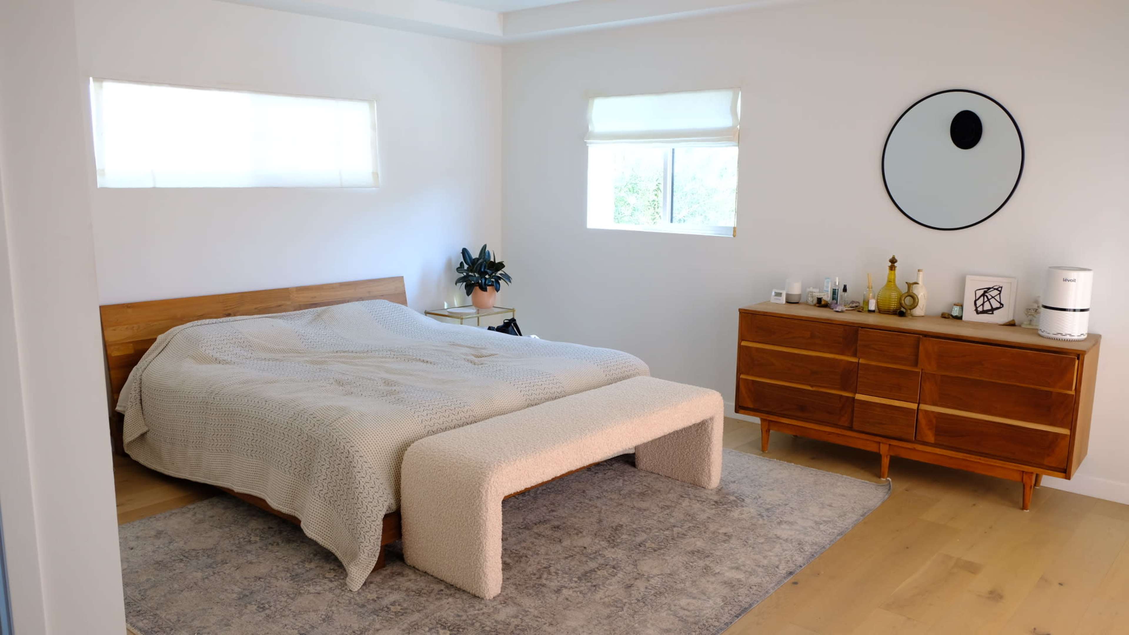 A minimalist bedroom with a wooden bed, a rug, a dresser, and a round mirror on the wall.