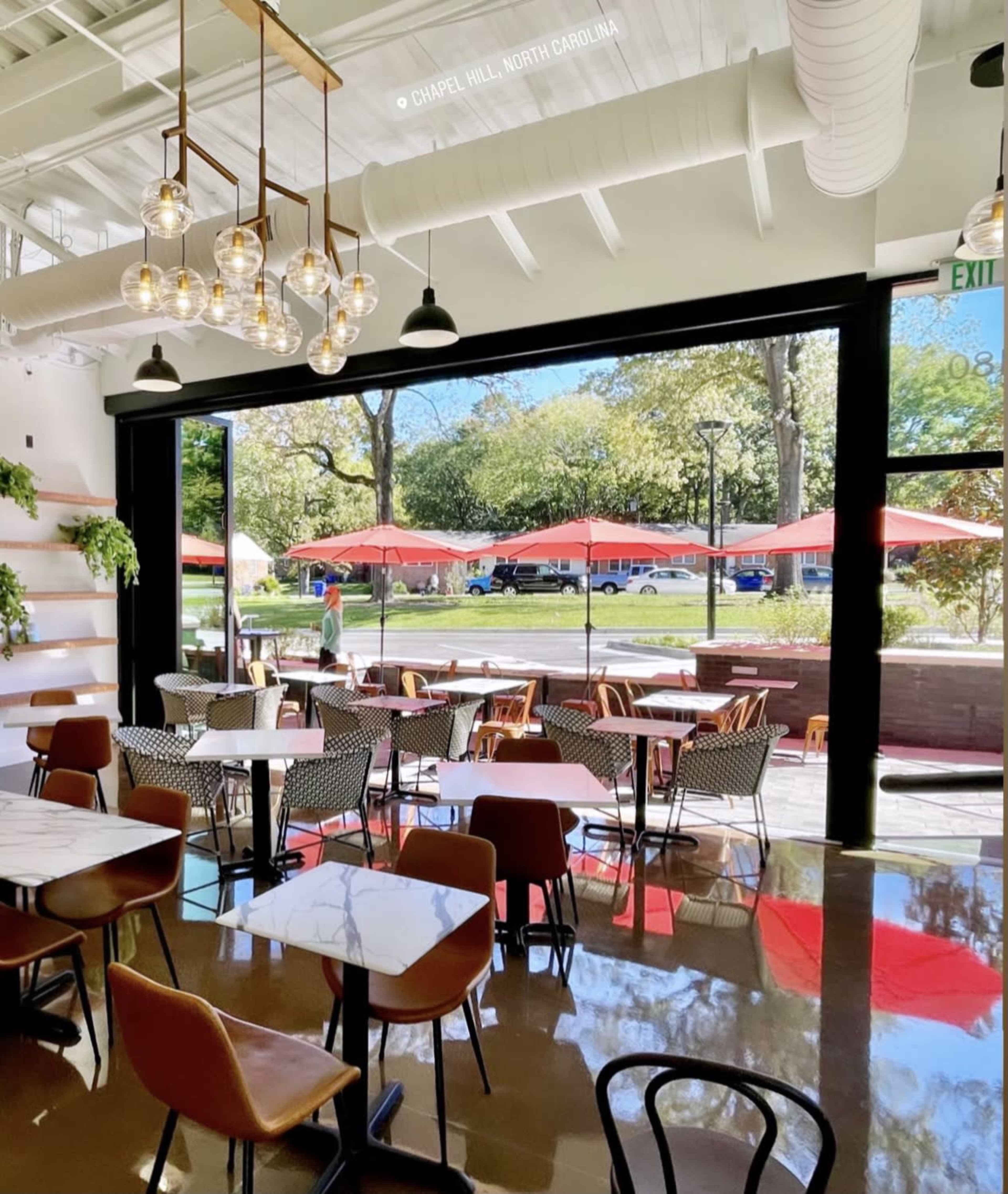 A modern café with indoor seating, large windows opening to an outdoor area with red umbrellas and greenery in Chapel Hill, North Carolina.