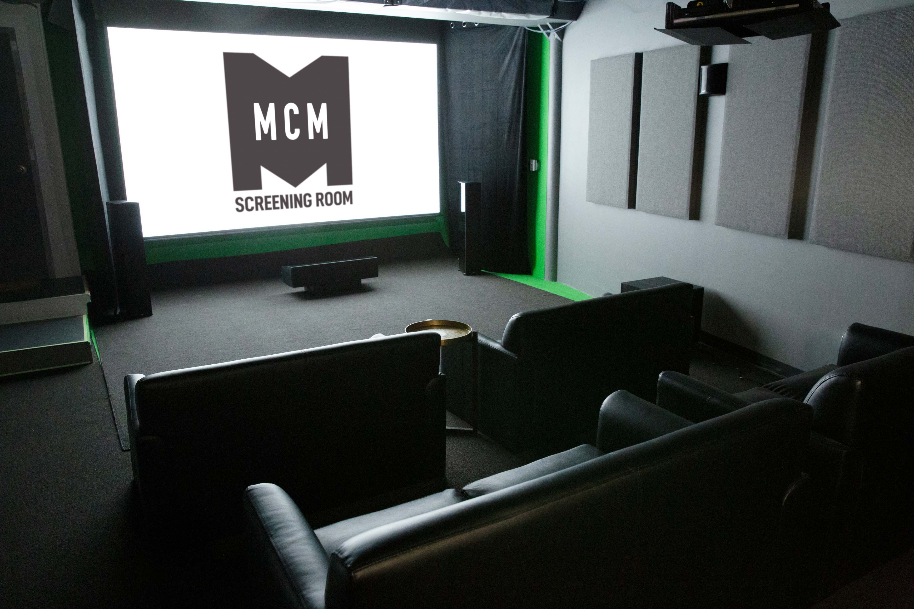 A dimly lit screening room with dark leather sofas facing a large screen displaying the logo "MCM SCREENING ROOM."
