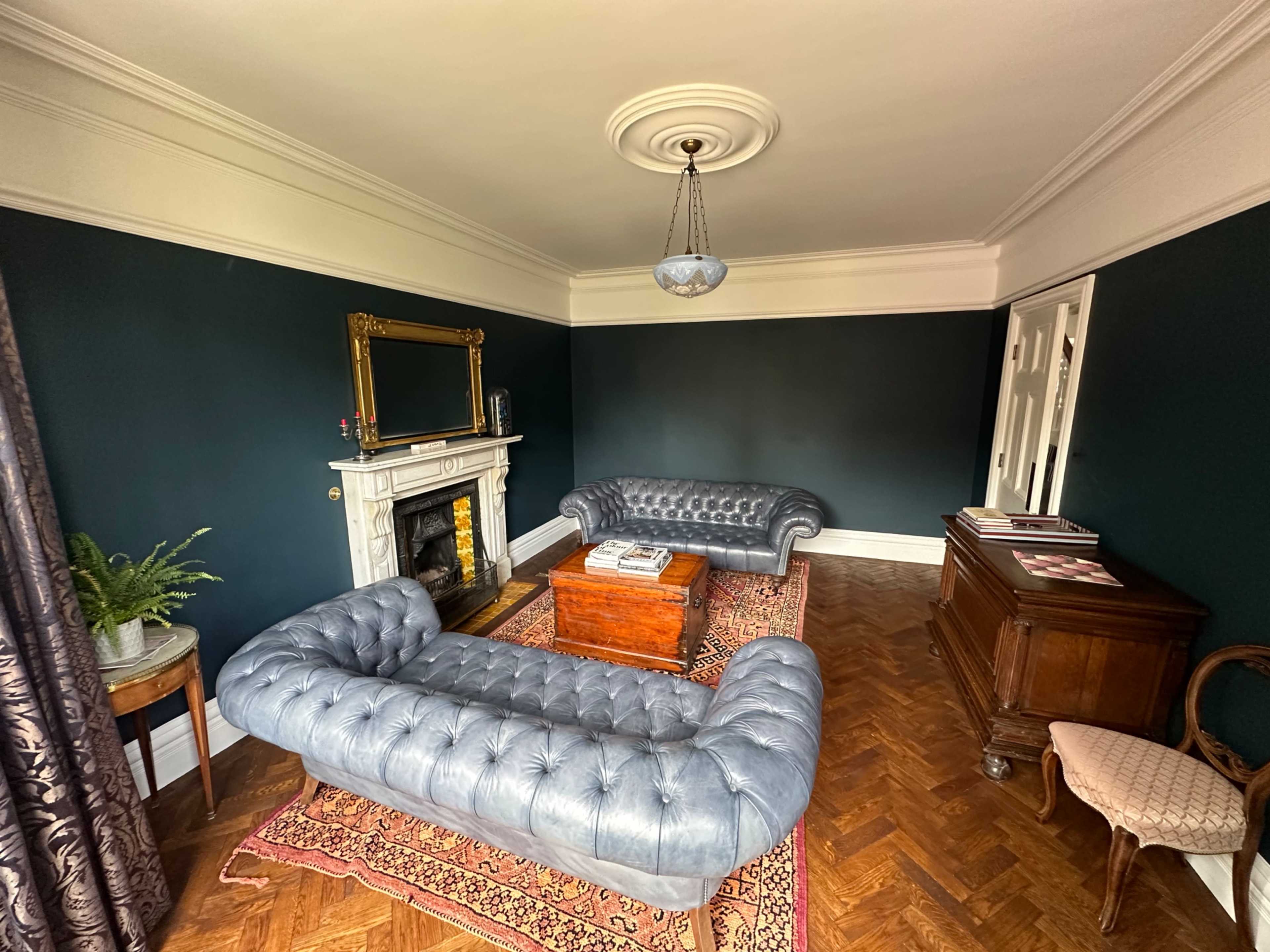 The room features two blue leather chesterfield sofas facing each other, a wooden coffee table in the center, and a decorative fireplace against a dark green wall.