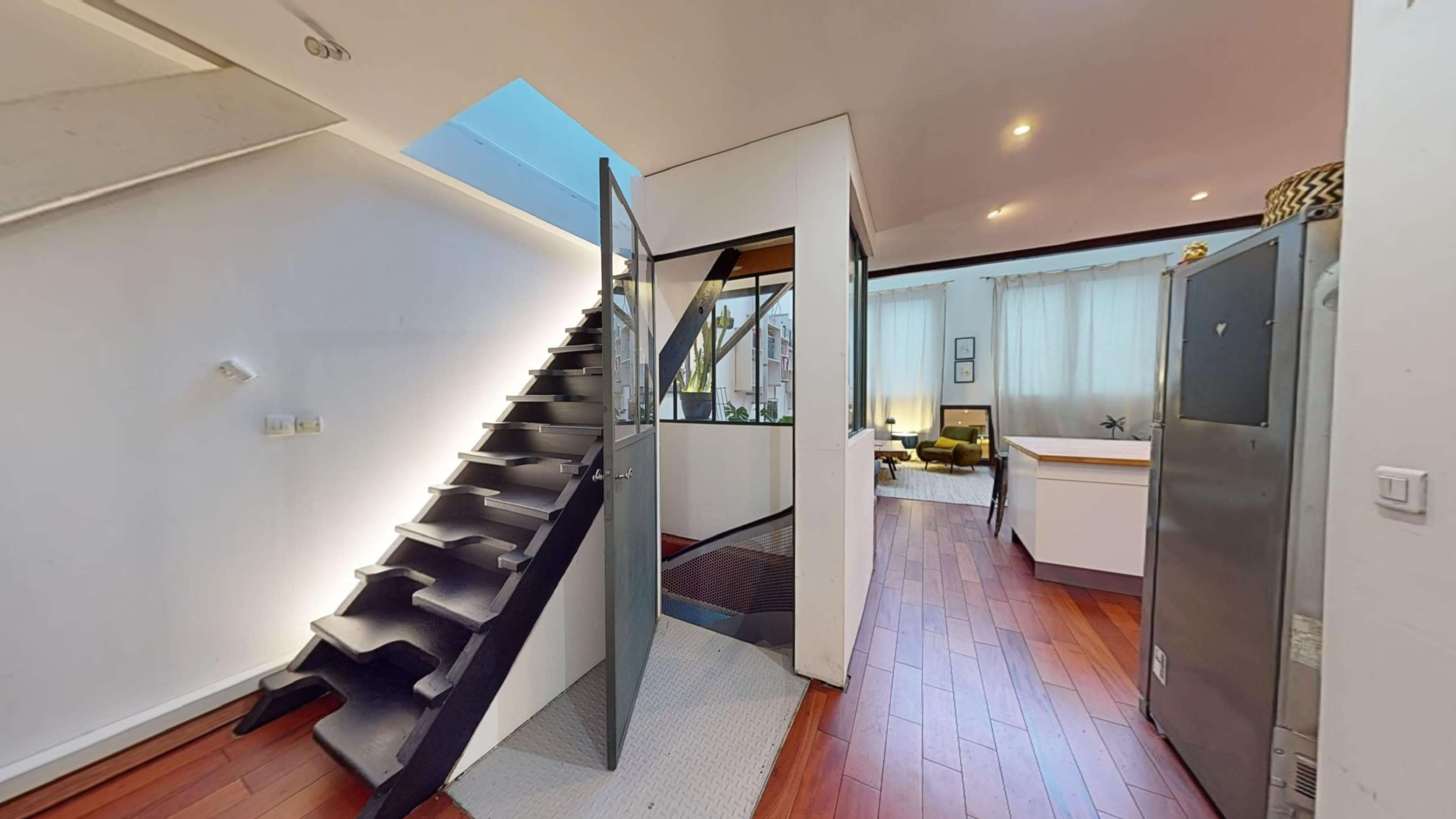 A modern interior space featuring a staircase leading to an upper level, with a view of a kitchen area and a living room in the background.