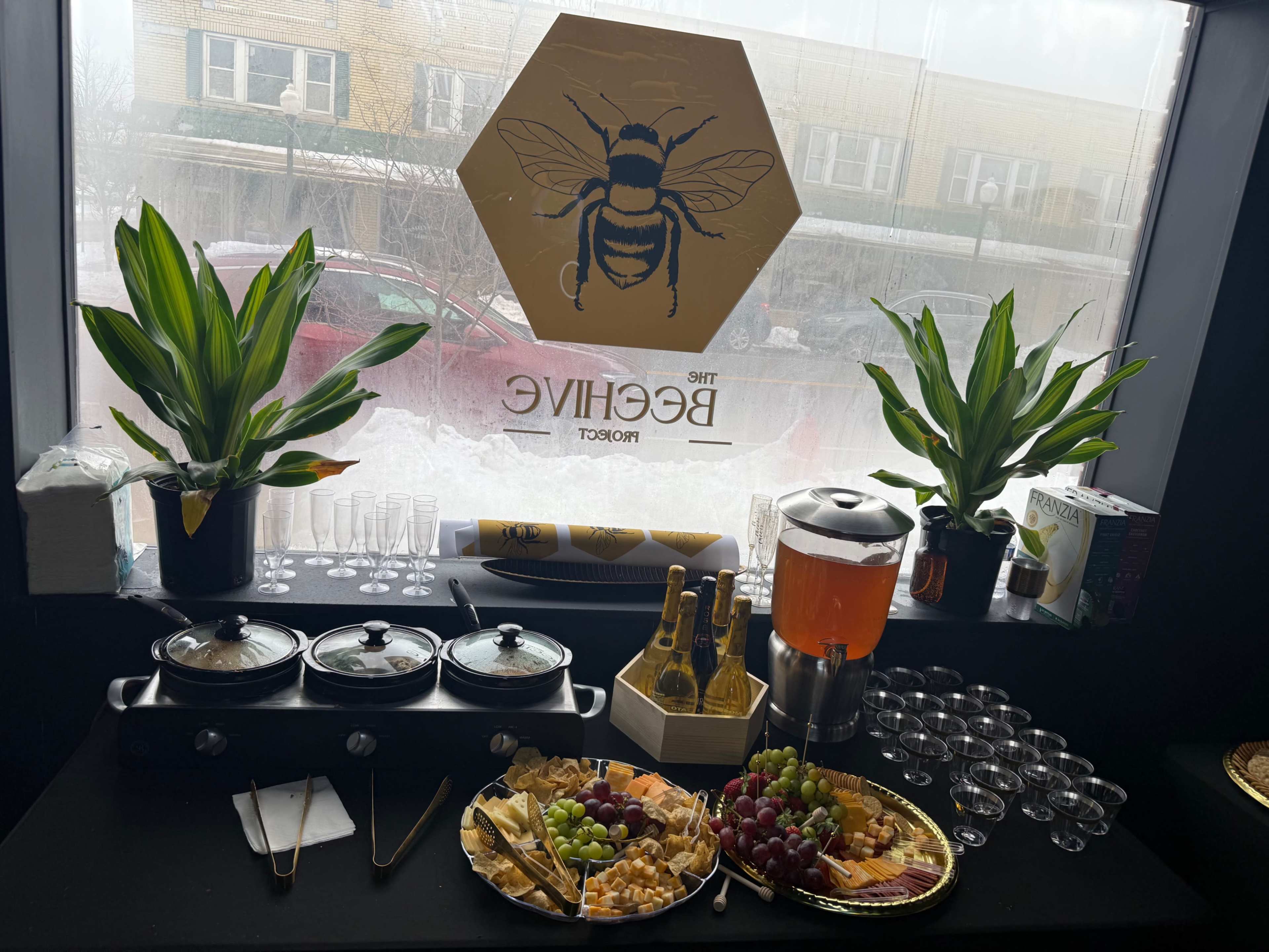 The image shows a buffet setup featuring a variety of cheeses, fruits, and beverages, with a bee-themed sign in the background.