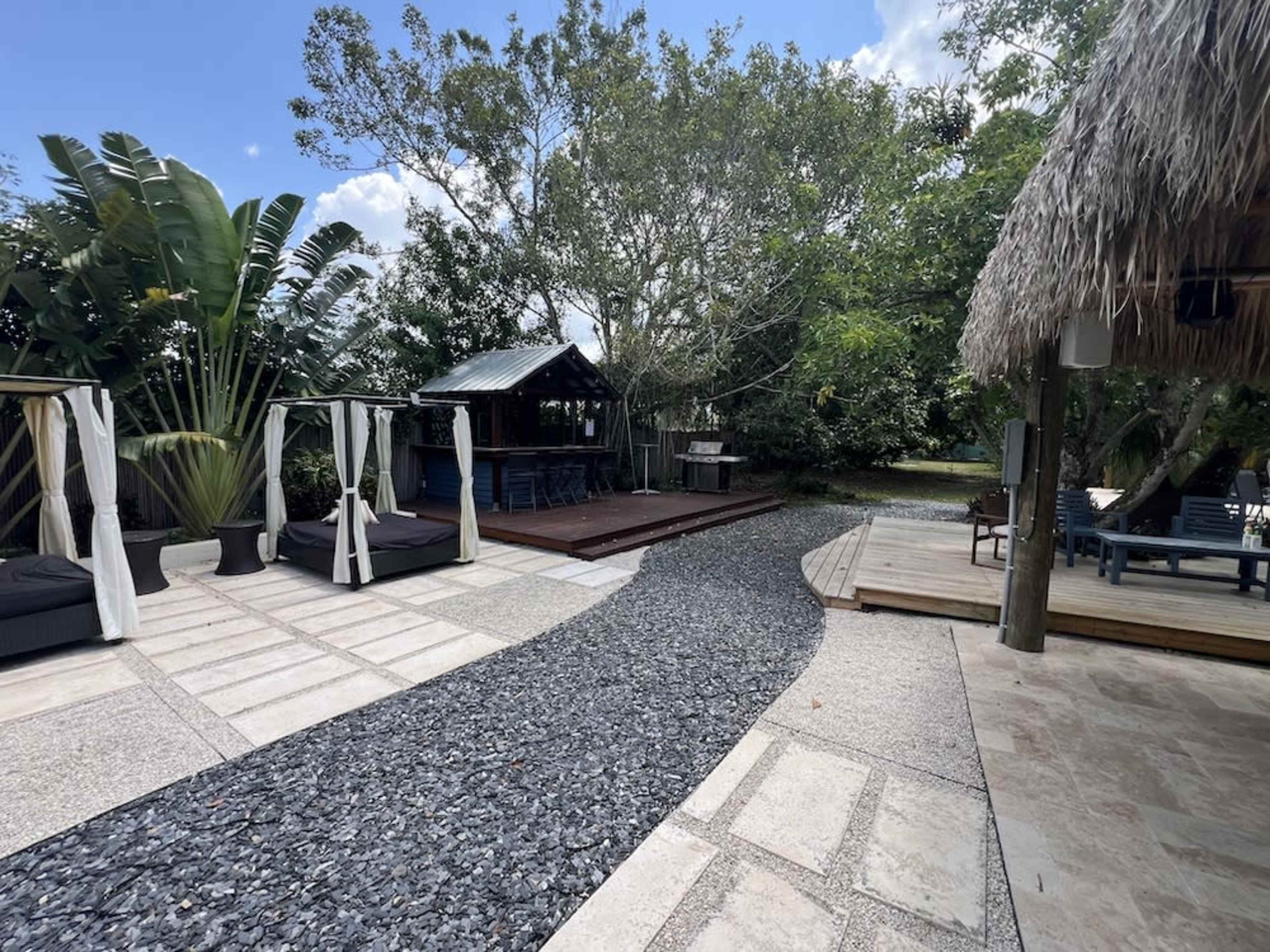 The image shows a landscaped outdoor area with a winding stone pathway leading to a shaded deck and seating areas surrounded by lush greenery.