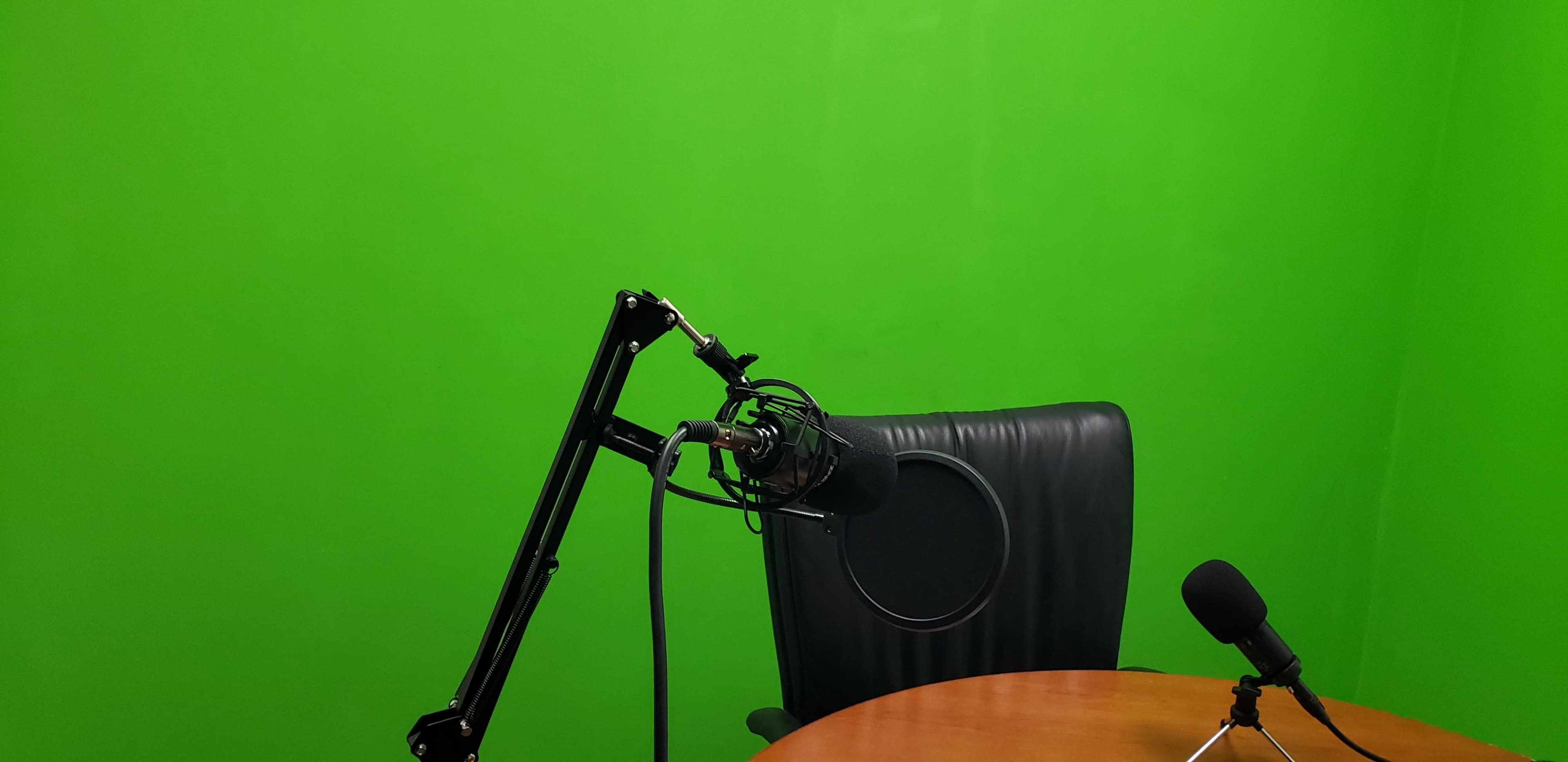A black microphone mounted on an adjustable arm next to a chair and a wooden table in front of a green screen.