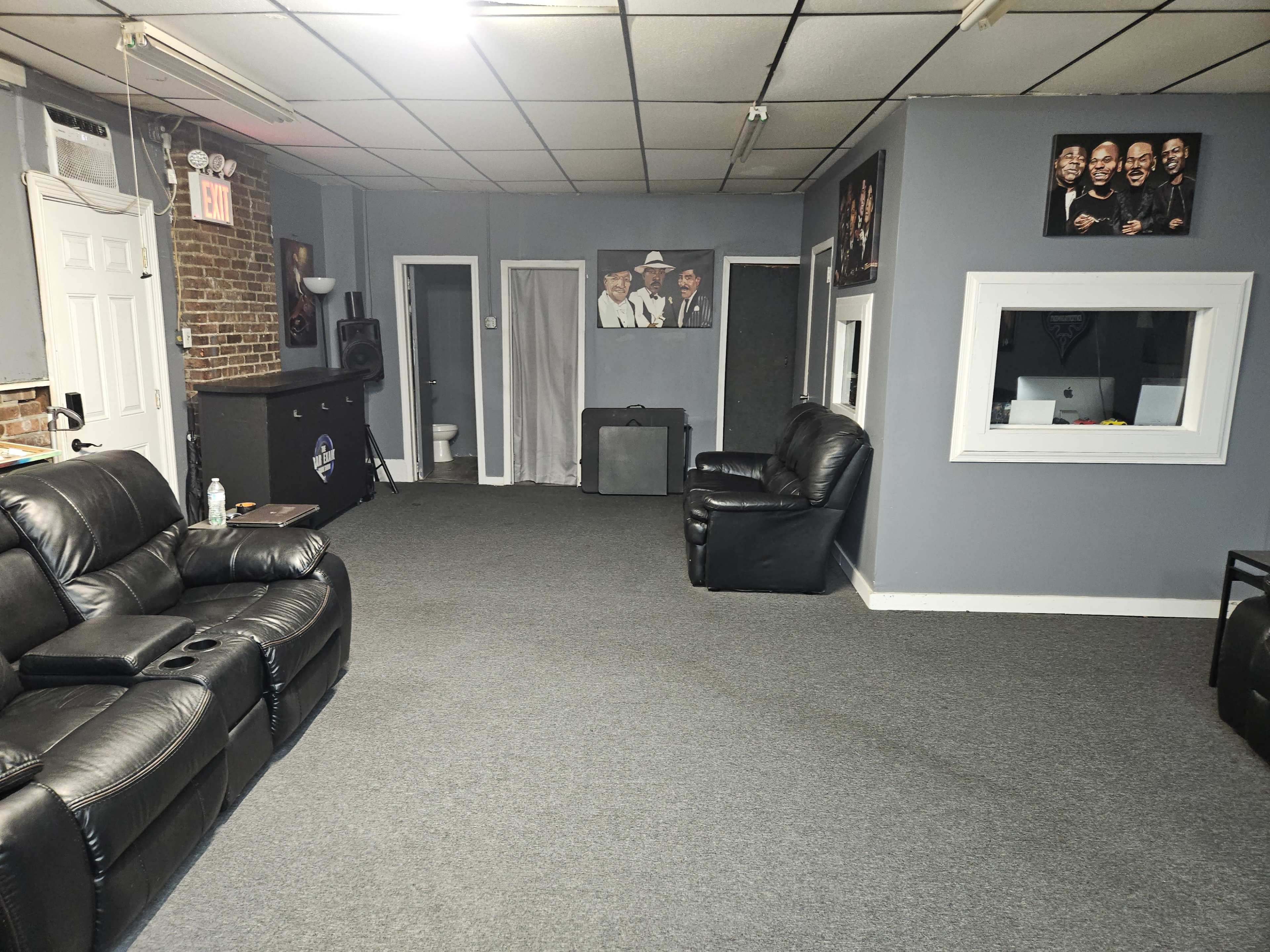 The image shows a spacious, gray-walled room with black leather seating, a bar counter, and framed photographs on the walls.