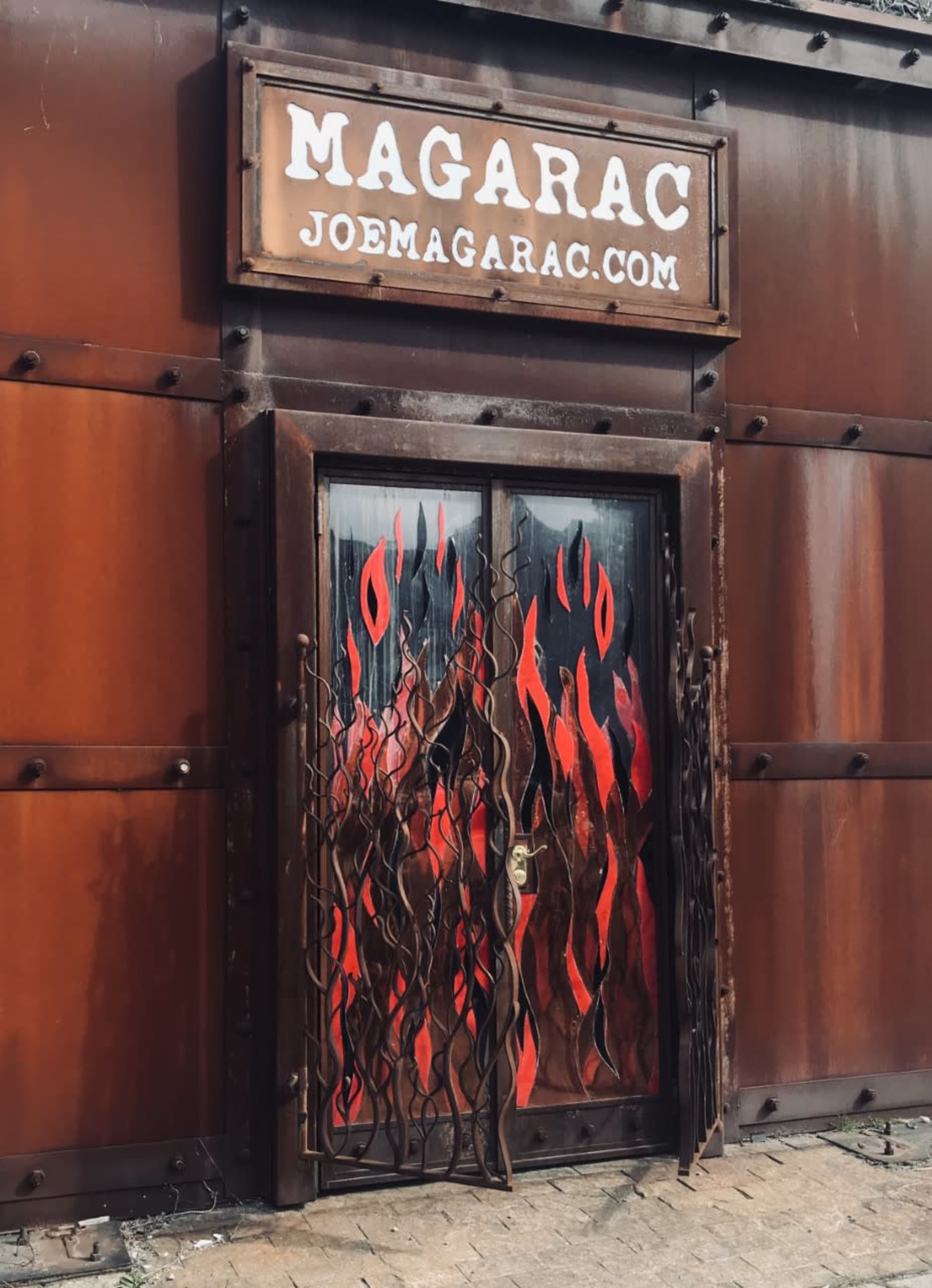 A metal door with a flame-themed design behind a rusted metal exterior and a sign overhead that reads "MAGARAC" along with the website "JOEMAGARAC.COM."