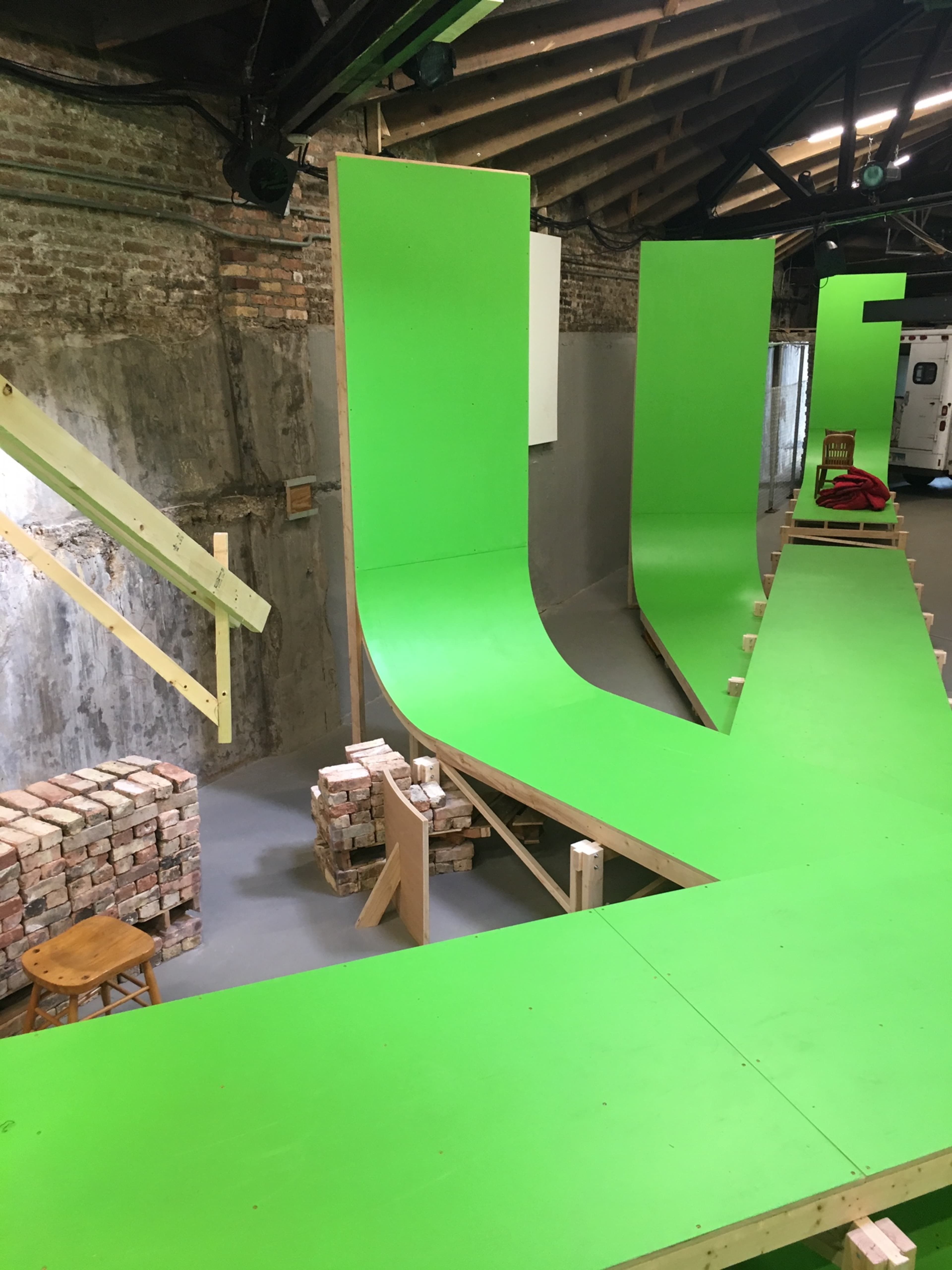 A large indoor ramp structure made of wood and painted bright green, with multiple curves and slopes, set against a brick wall in an industrial space.