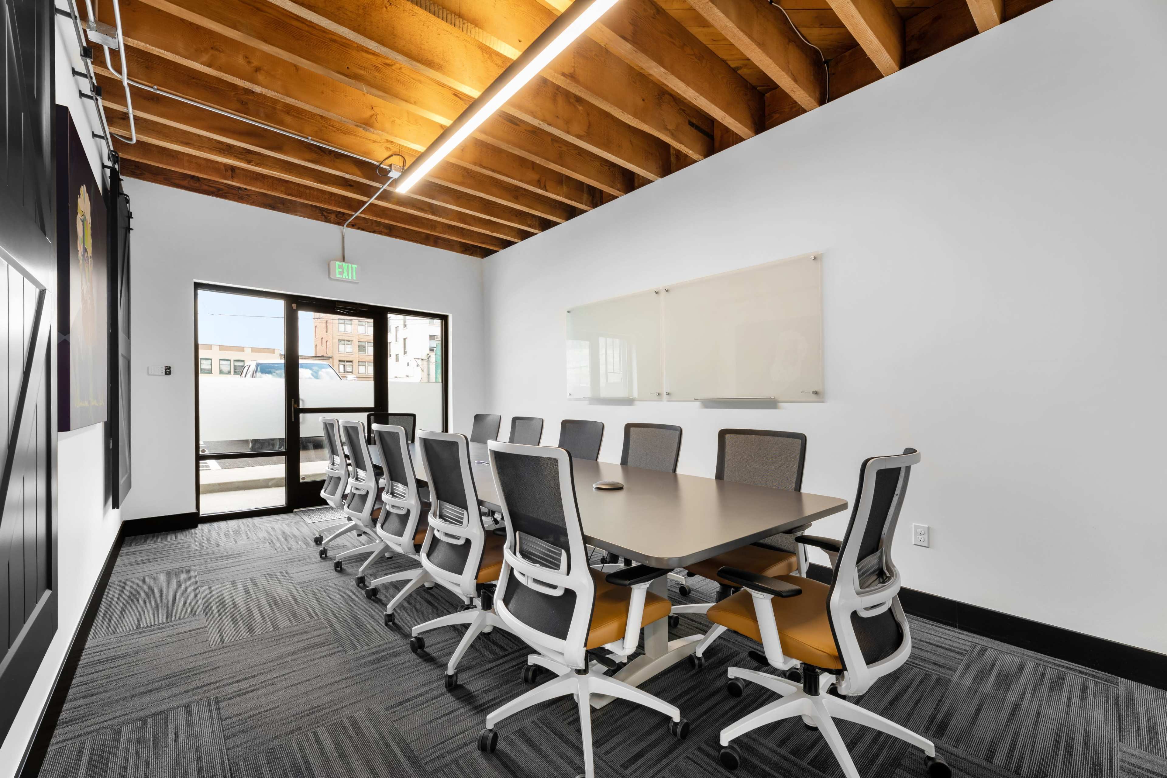 A large conference room features a long table surrounded by office chairs, with a glass door leading to an outdoor area and exposed wooden beams overhead.