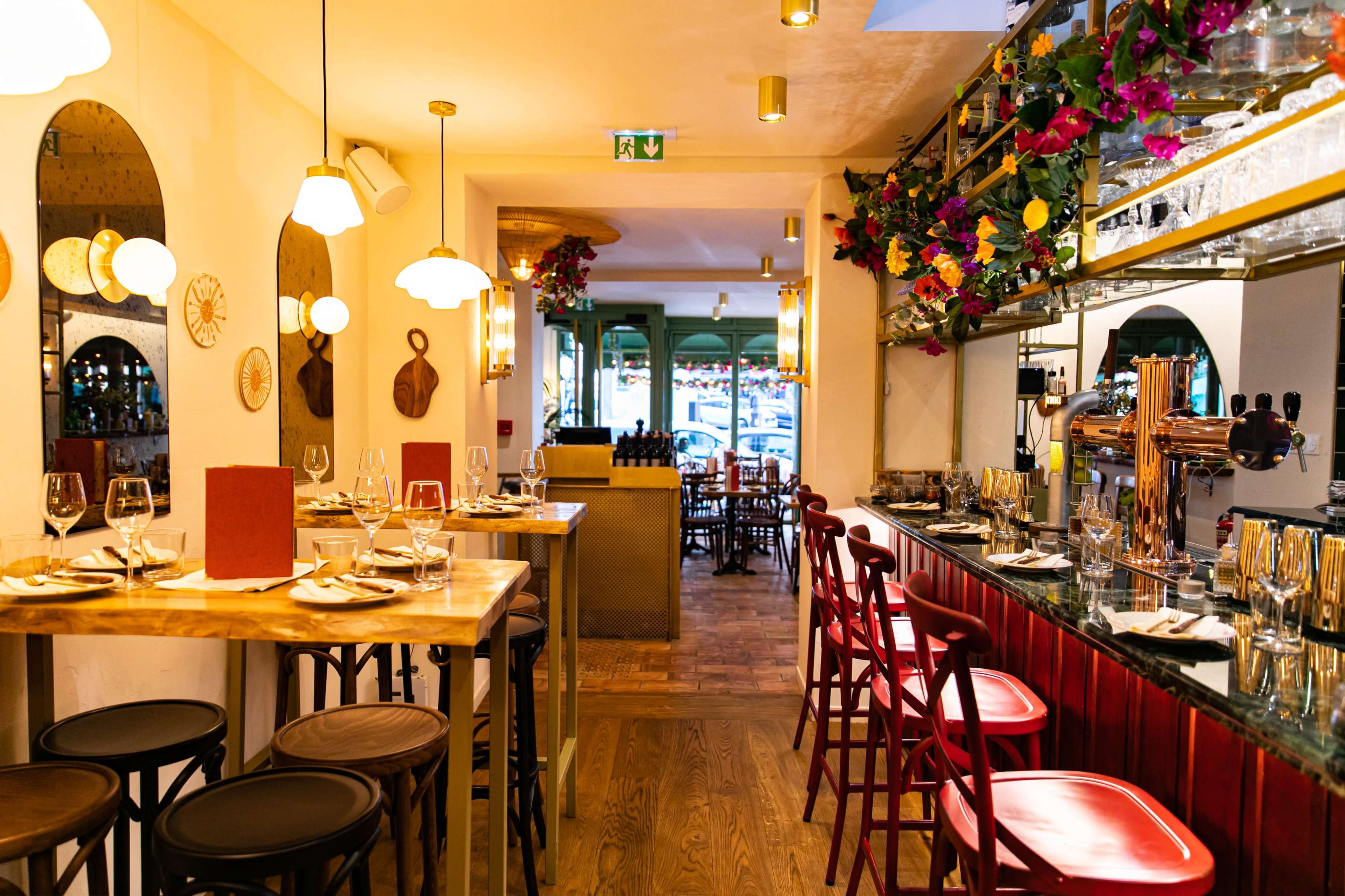 A brightly lit restaurant interior with wooden tables, bar stools, and a decorated bar area featuring hanging plants and a mirrored wall.