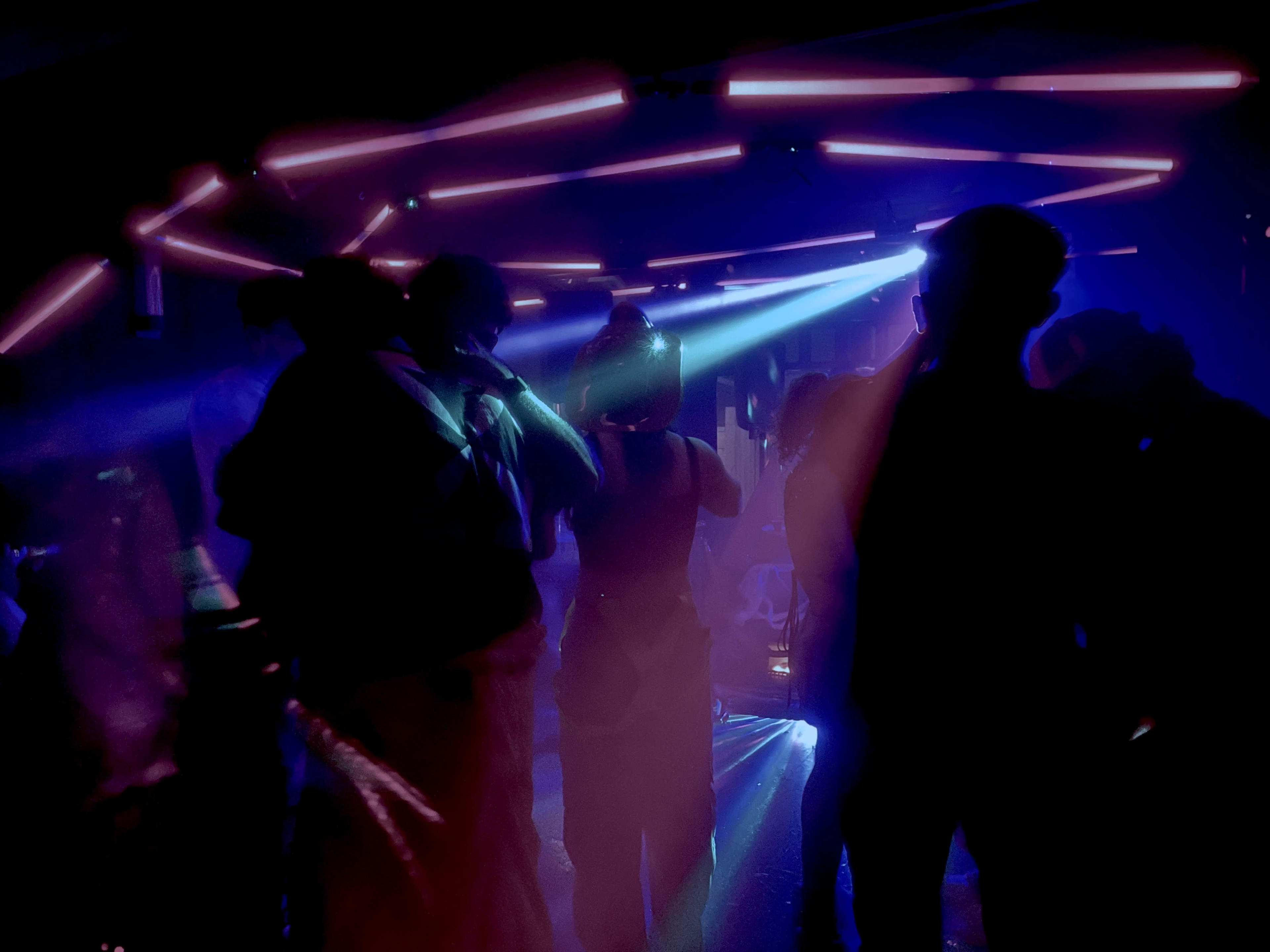 A group of people dance in a dimly lit room illuminated by colorful neon lights and beams of smoke.