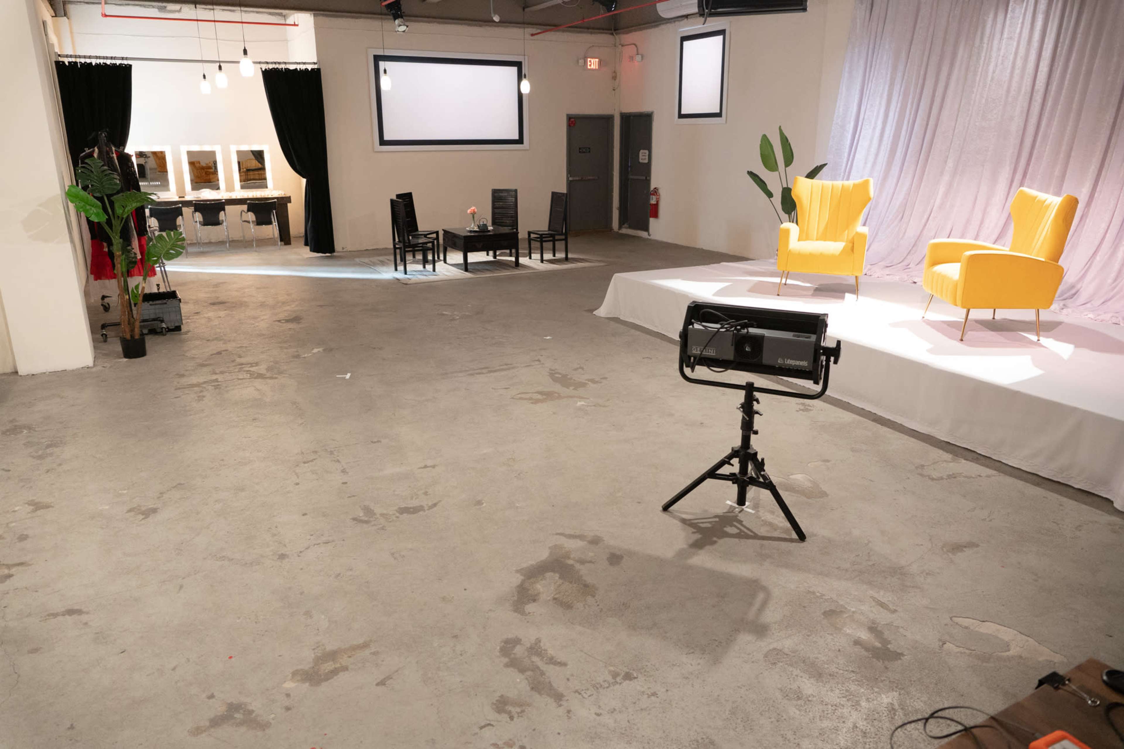 A spacious room with two yellow chairs on a raised platform, positioned across from a seating area with a small table and four black chairs, and a row of mirrors along one wall.