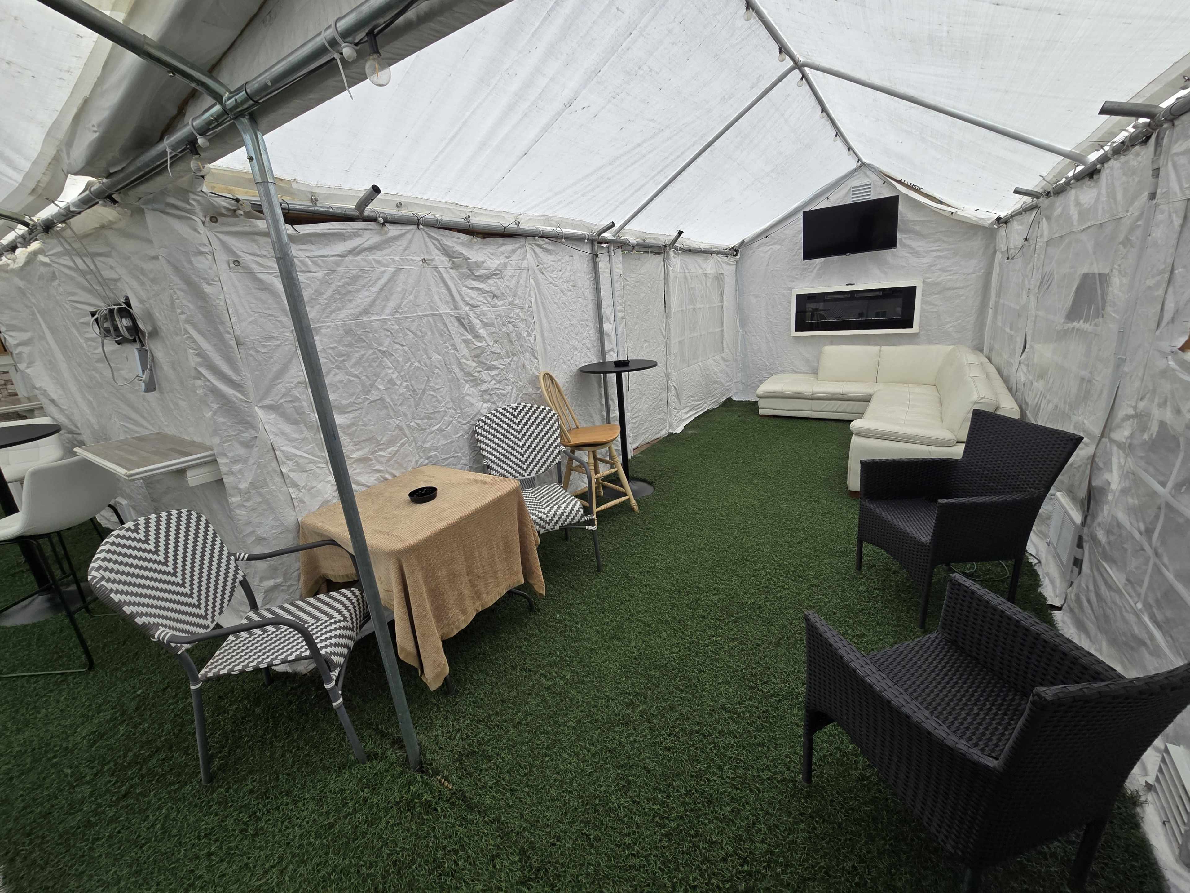 The image shows a tented space with artificial grass flooring, furnished with a white sectional sofa, several chairs, a small table, and a television mounted on the wall.