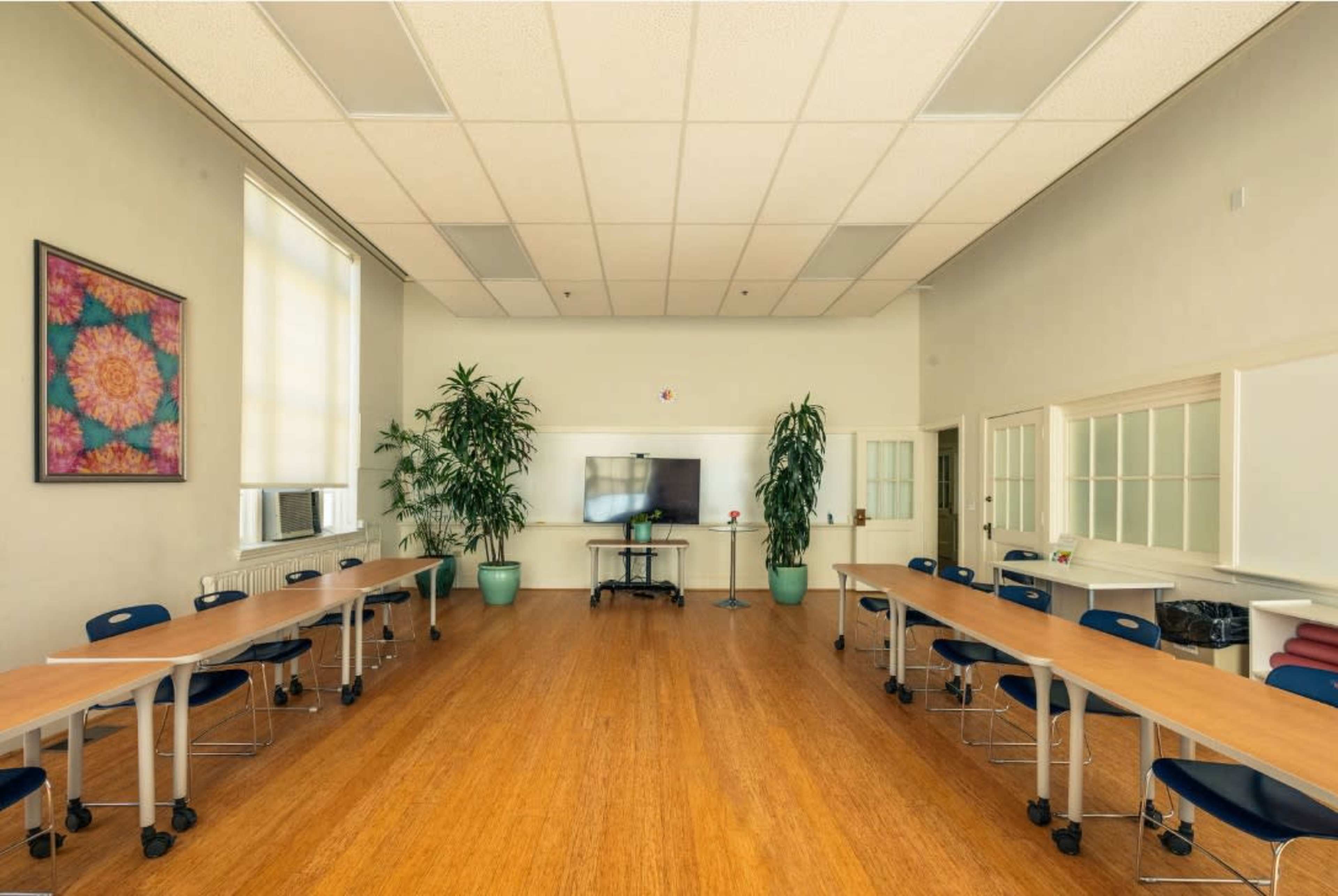 A spacious classroom features two rows of tables and chairs, large windows, and potted plants along the walls.