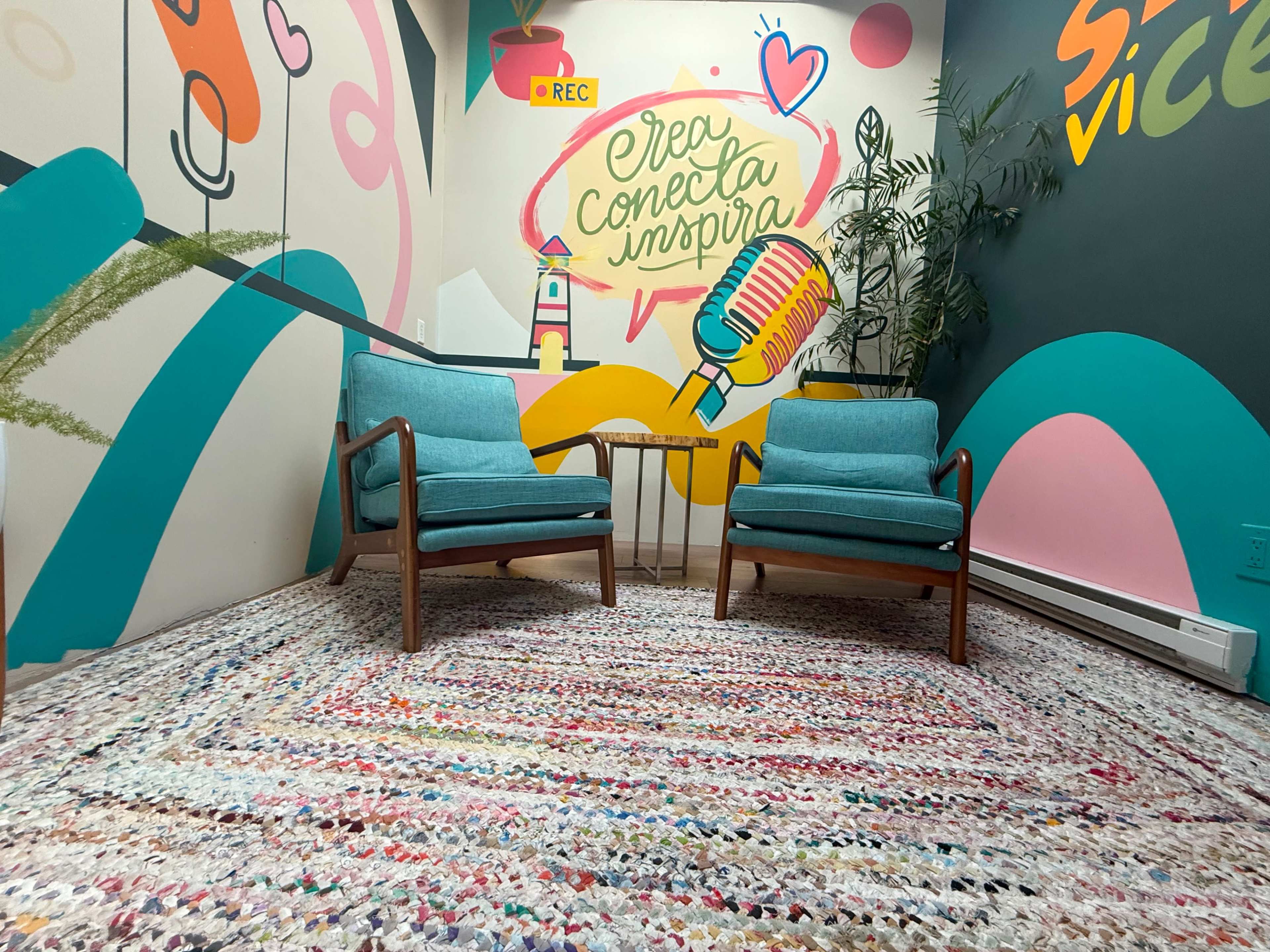 The image shows a brightly colored room featuring two teal armchairs on a textured rug, with a vibrant mural on the wall that includes a microphone and various graphic designs.