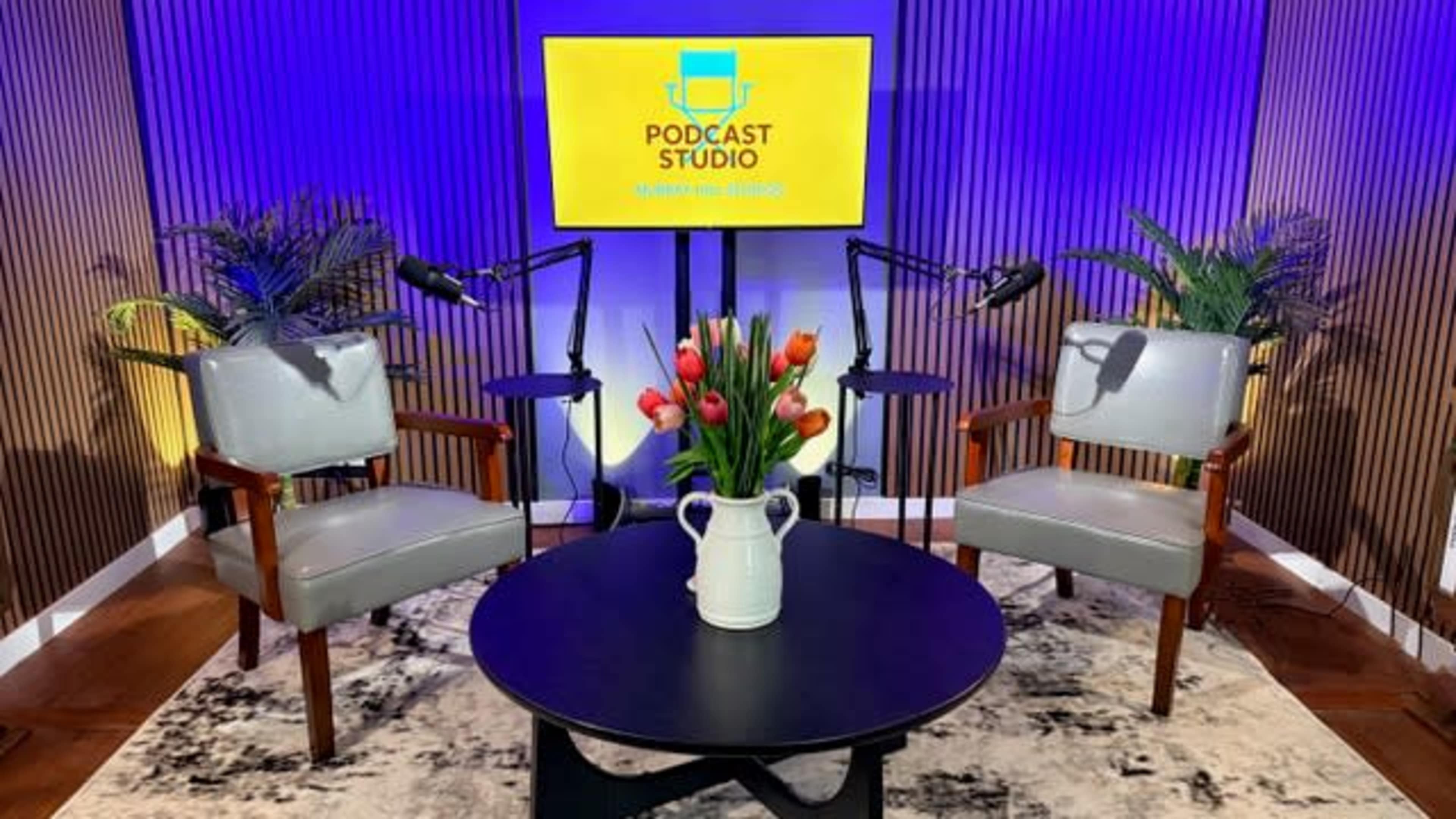 The image shows a modern podcast studio setup with two chairs, a black circular table, and a screen displaying the text "PODCAST STUDIO" along with microphones and decorative plants.