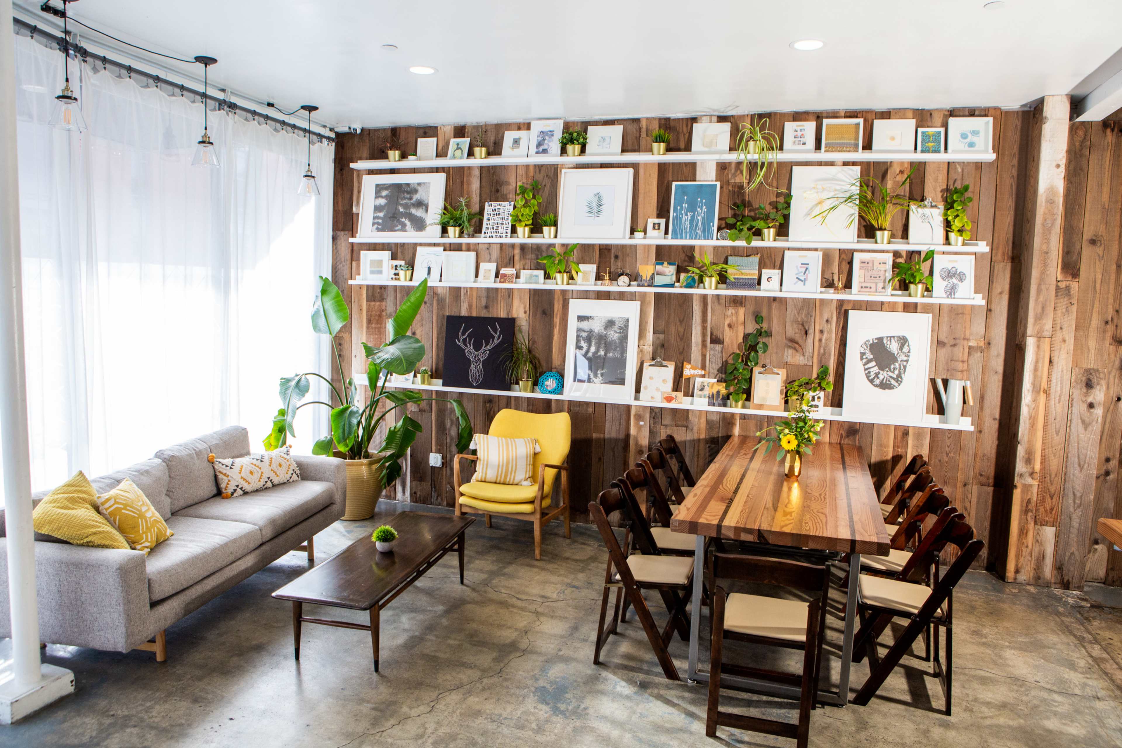 The image shows a cozy interior space featuring a wooden wall with shelves displaying framed art and plants, alongside a gray couch, a yellow chair, and a dining table with chairs.