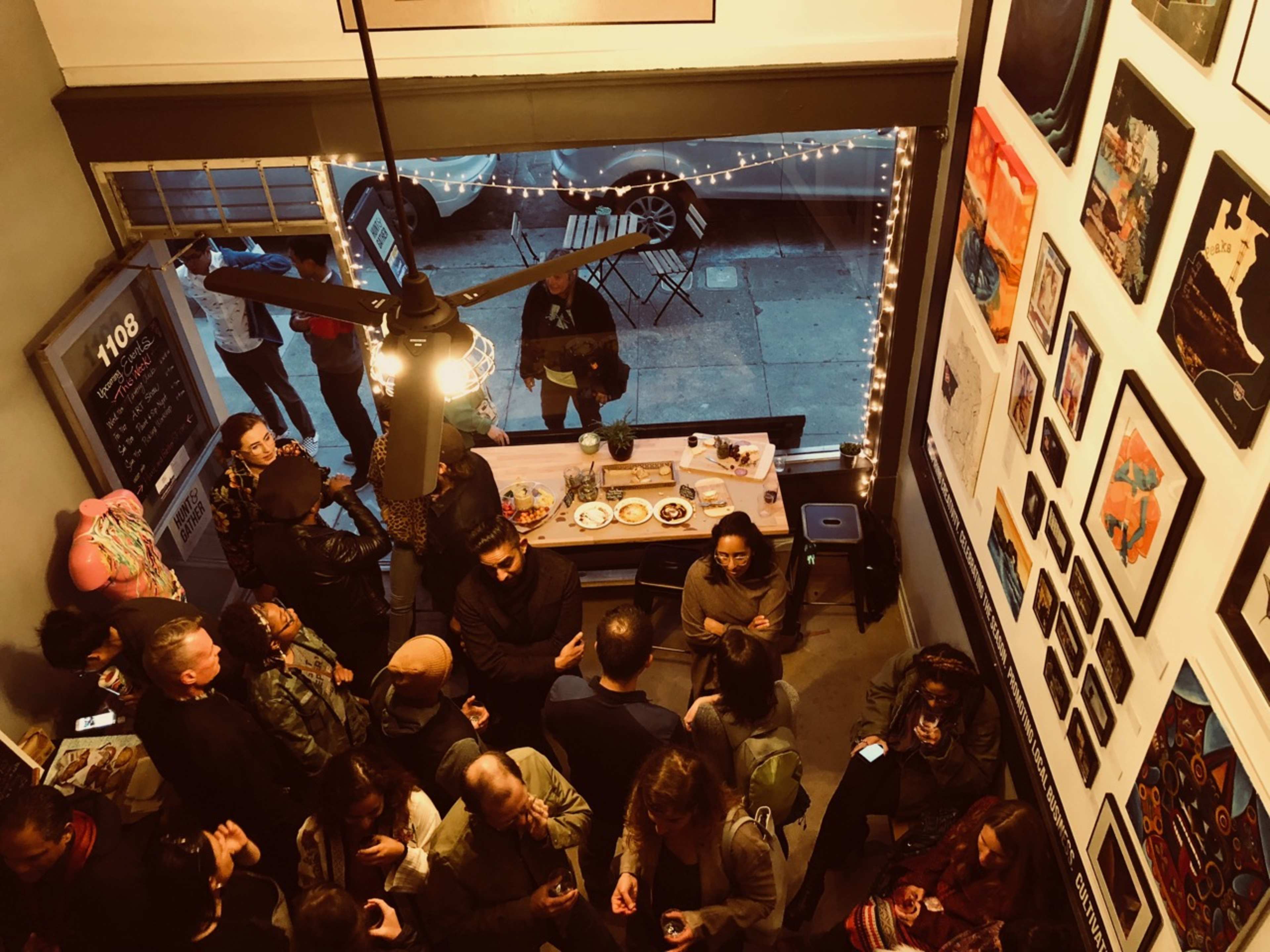 A crowded room with people socializing around a table filled with food and drinks, while art is displayed on the walls and lights hang above.