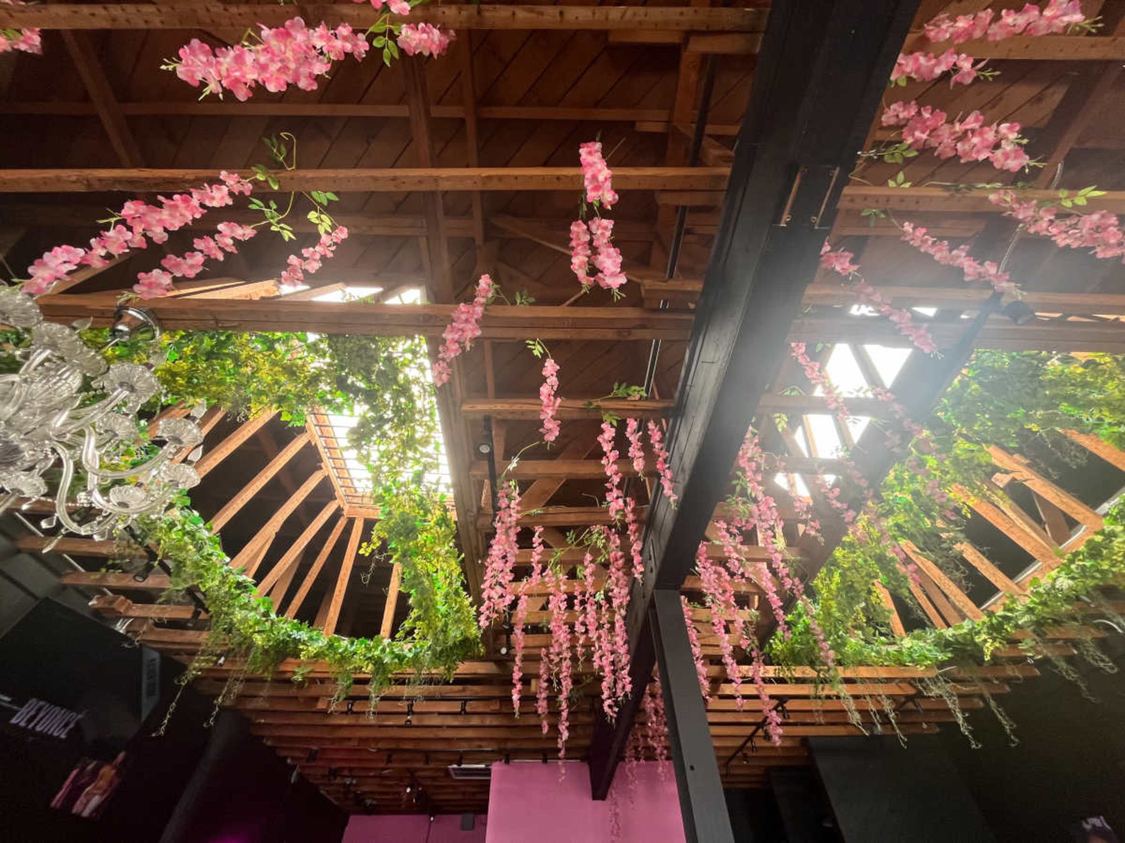 The image shows a ceiling adorned with hanging pink flowers and greenery, framed by wooden beams and skylights.