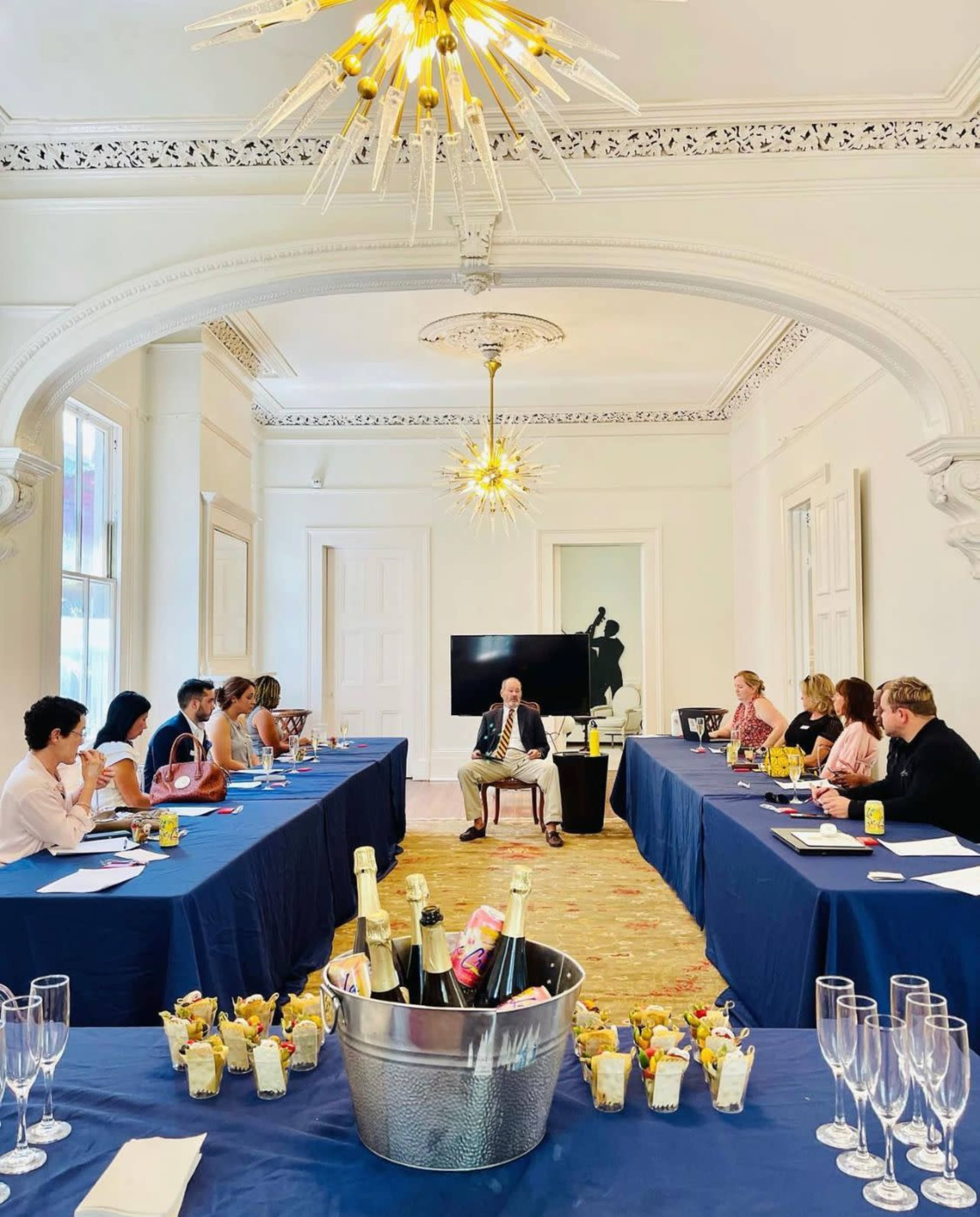 A group of people is seated around two rectangular tables in a bright, elegantly decorated room, with a man in a suit speaking to them, while drinks and snacks are arranged in the center.