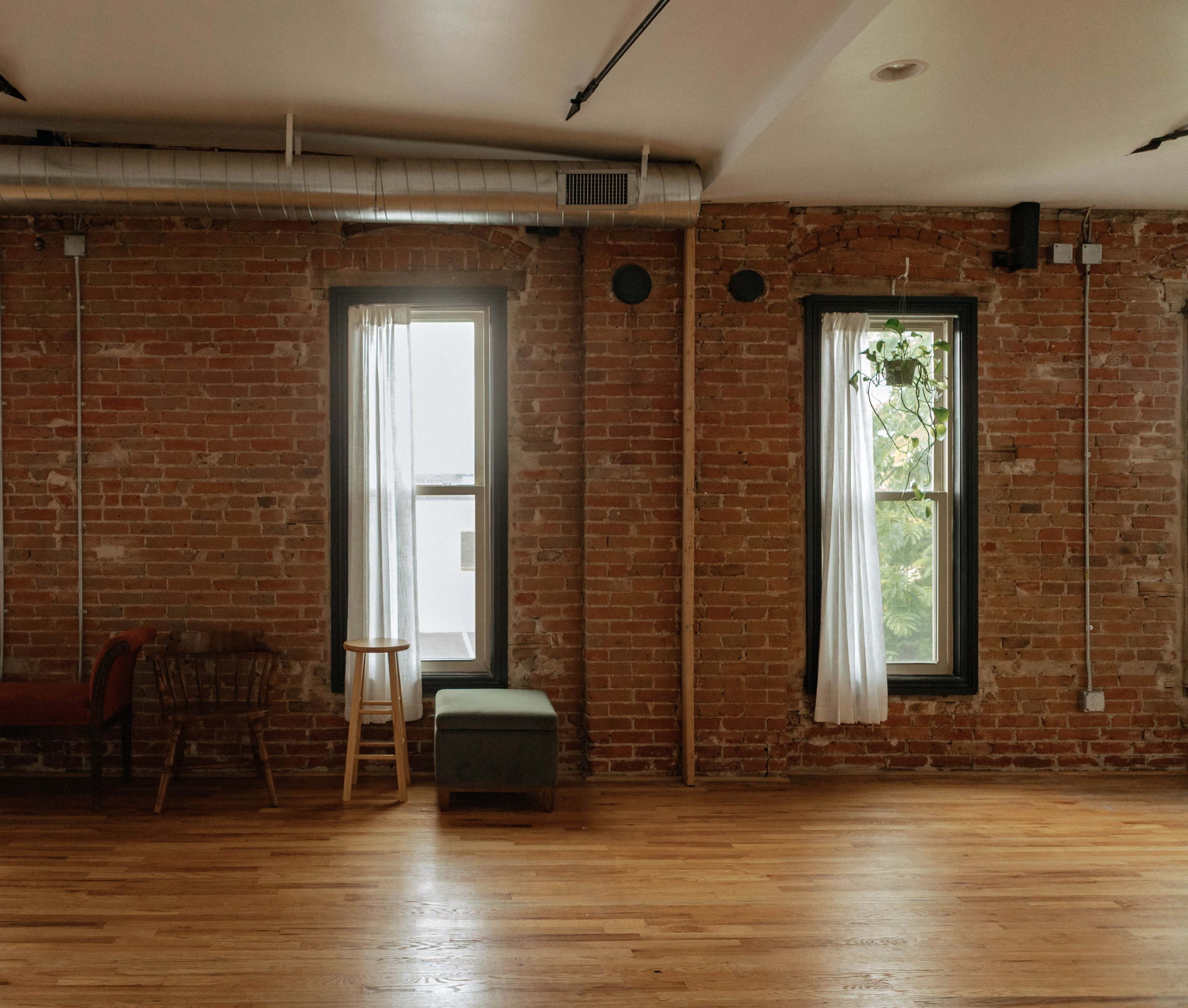 The room features exposed brick walls, large windows with sheer white curtains, a wooden floor, and minimal furnishings including a stool and a bench.