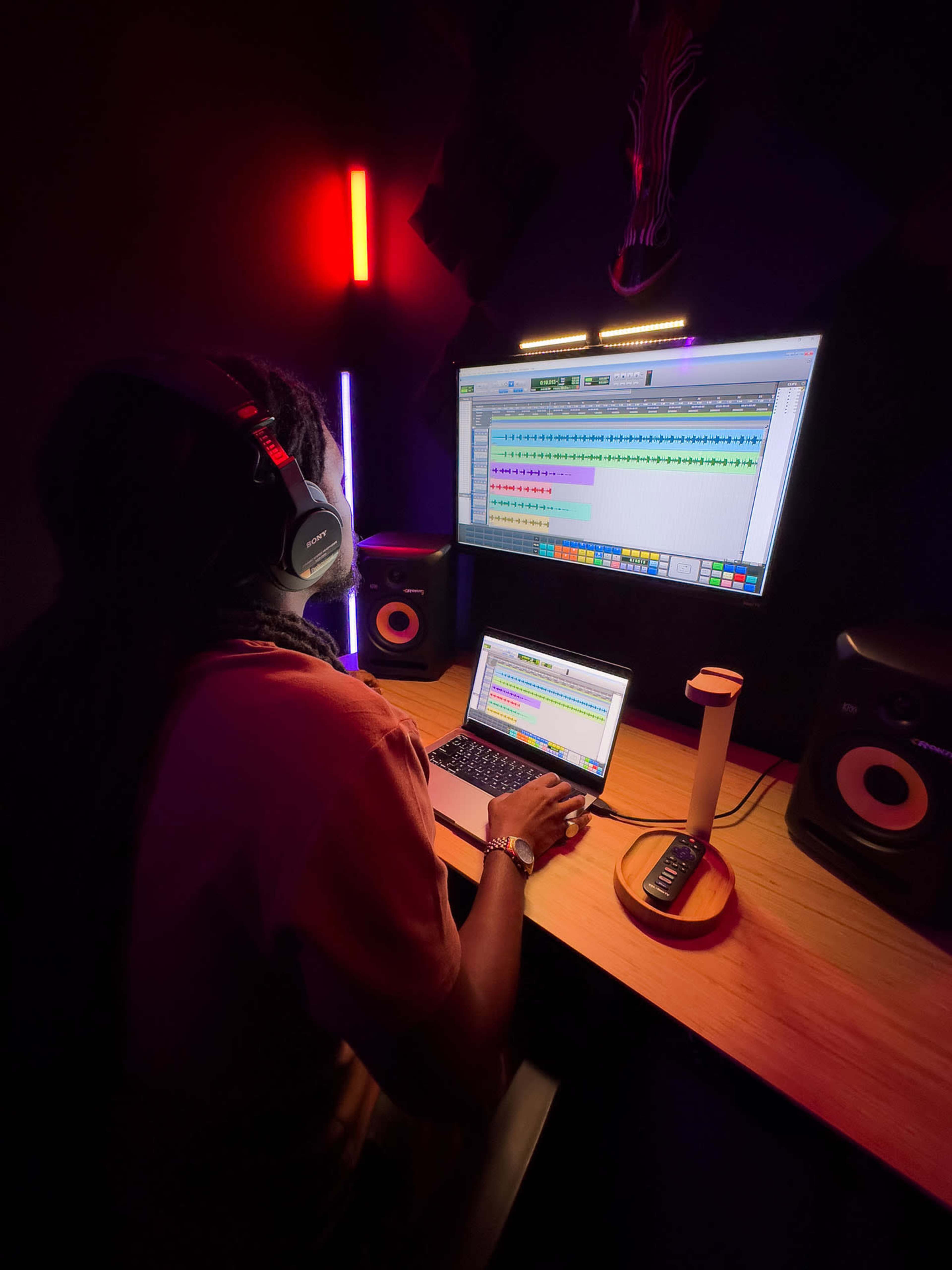 A person is seated at a desk in a dark room, using a laptop and headphones while working on music production software, with illuminated monitors and speakers visible.