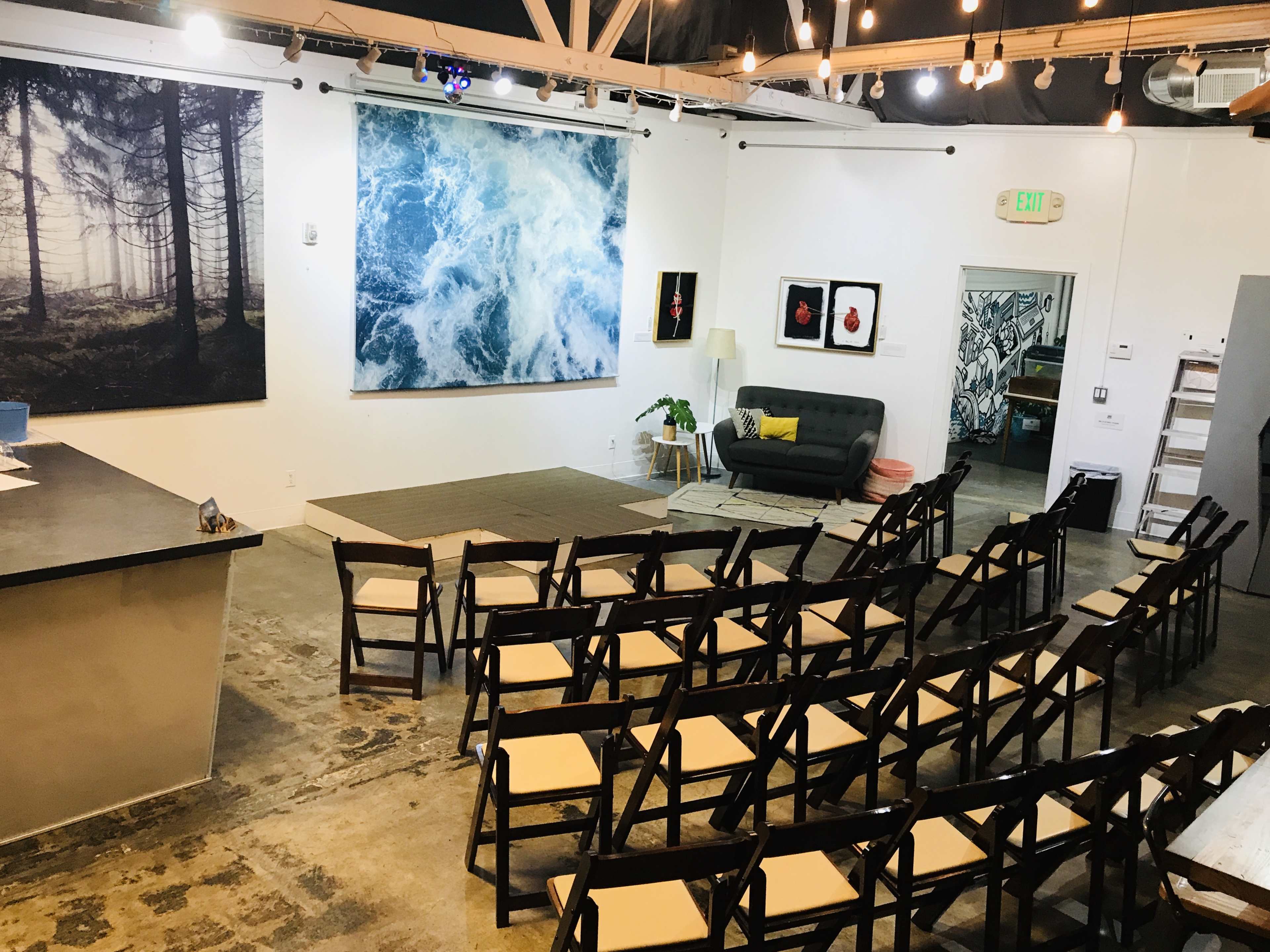 The image shows a spacious interior room with arranged rows of folding chairs facing a small stage area, surrounded by various art pieces on the walls.