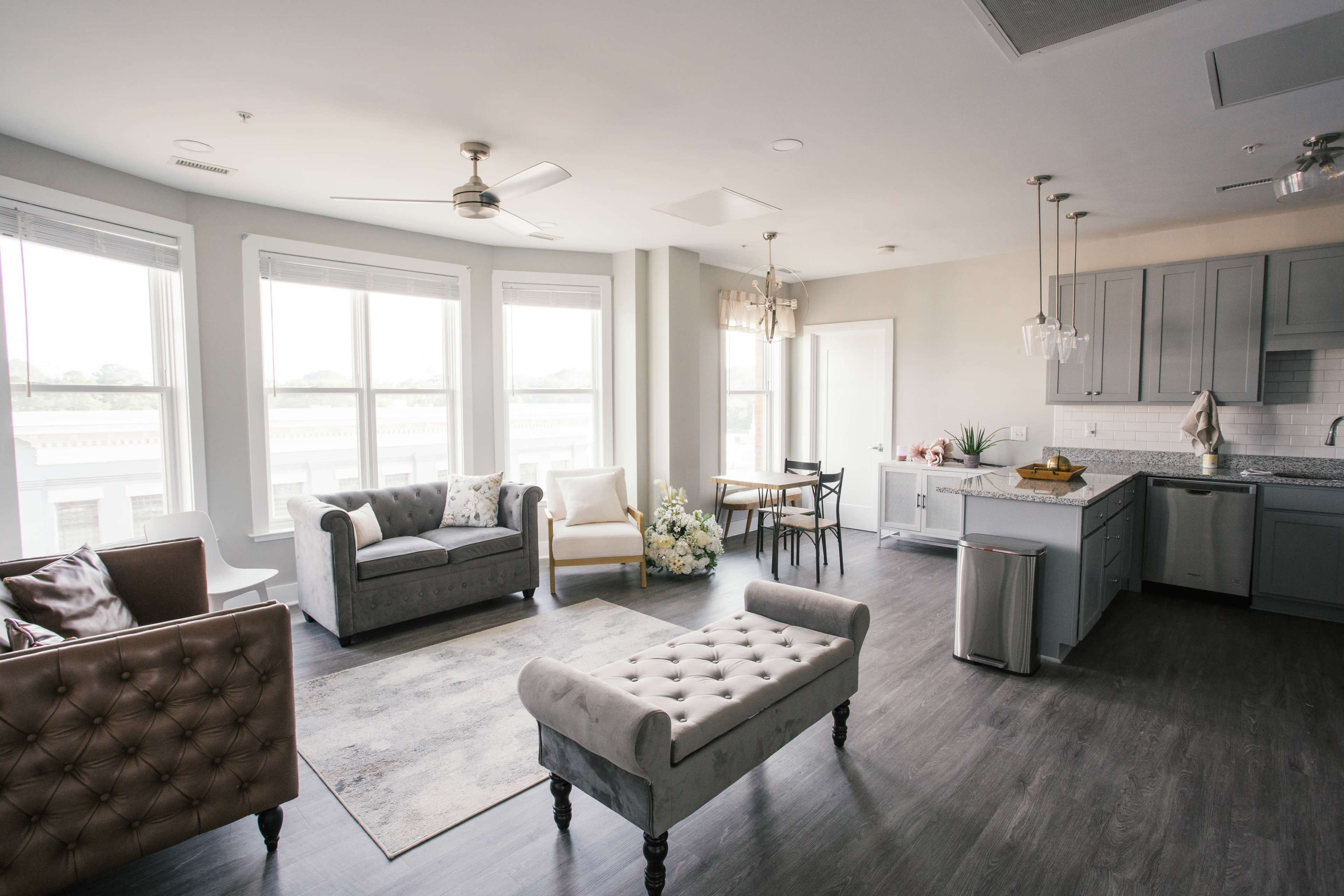 The image shows a modern, open-concept living area with large windows, a sectional sofa, and a kitchen featuring gray cabinetry and a central island.