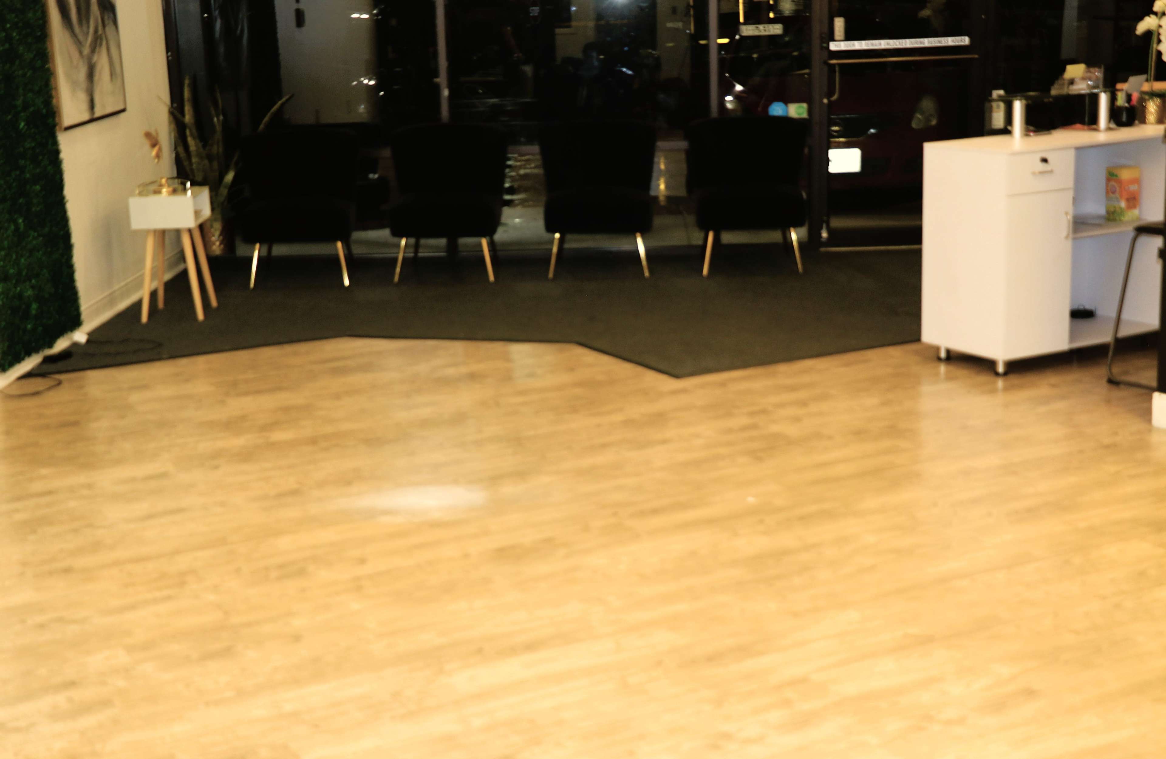 The image shows a minimalist interior space with a light-colored wooden floor, a black carpet section, and four black chairs arranged near a large front window.