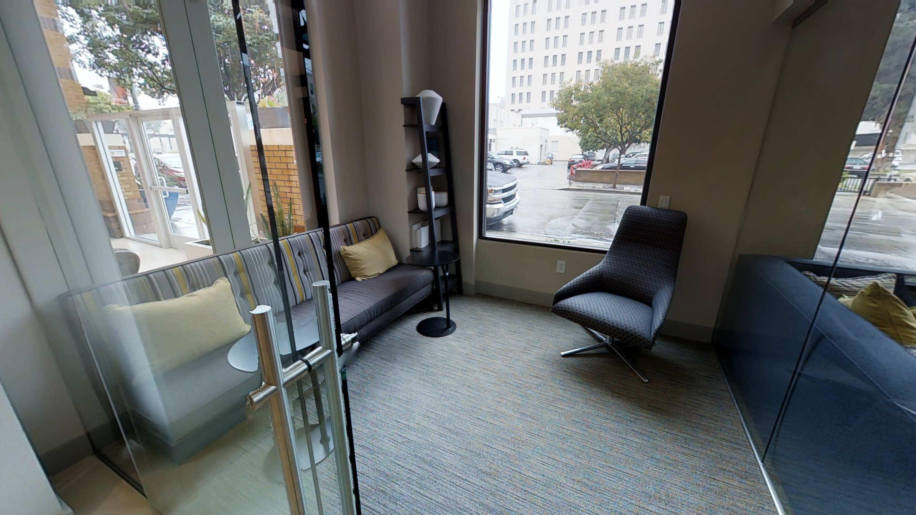 The image shows a small lounge area with a couch, a single chair, and a bookshelf, all situated next to large windows.