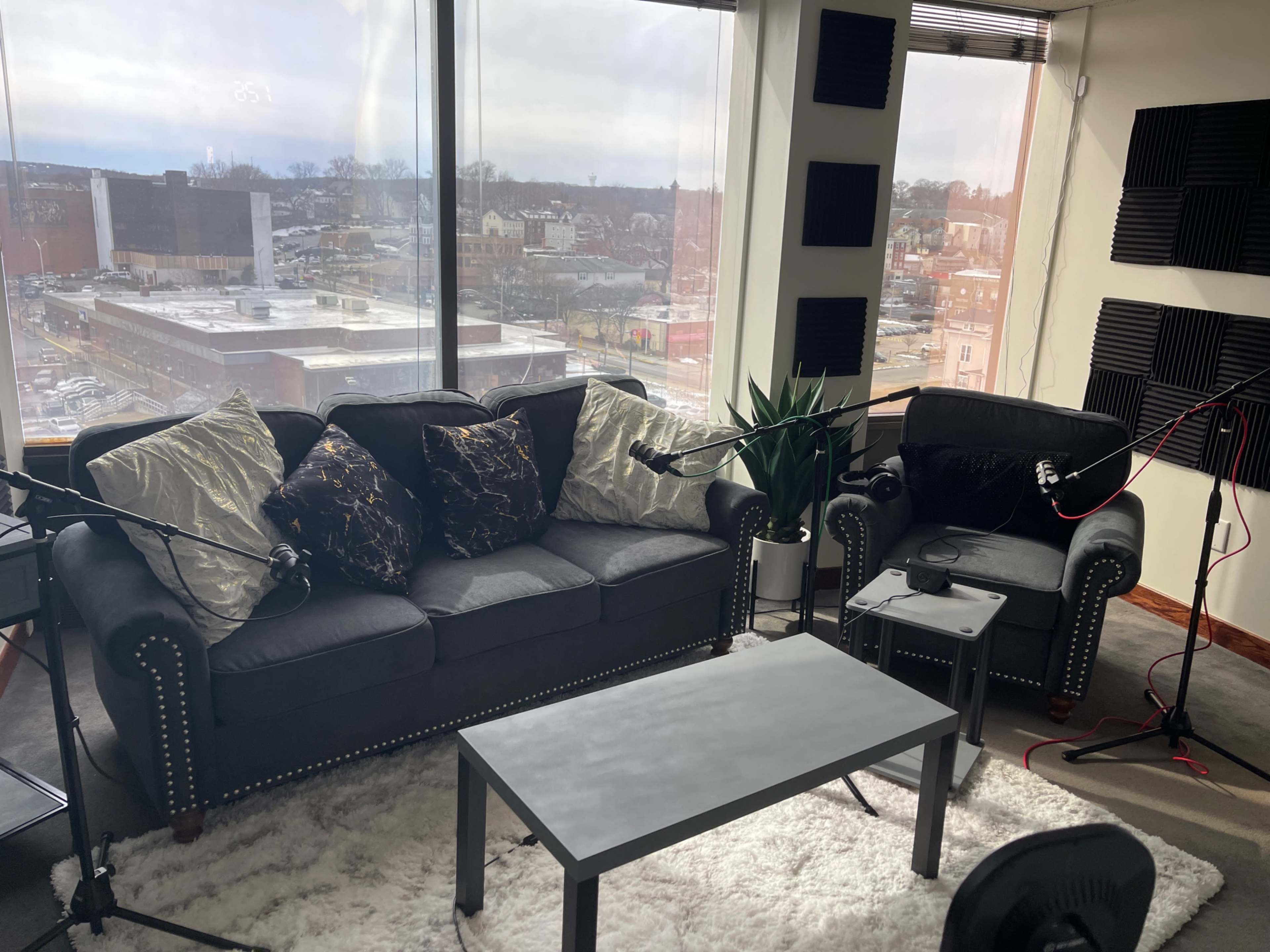 A modern recording studio with a black sofa, a matching chair, a coffee table, and audio equipment arranged around a large window overlooking an urban landscape.