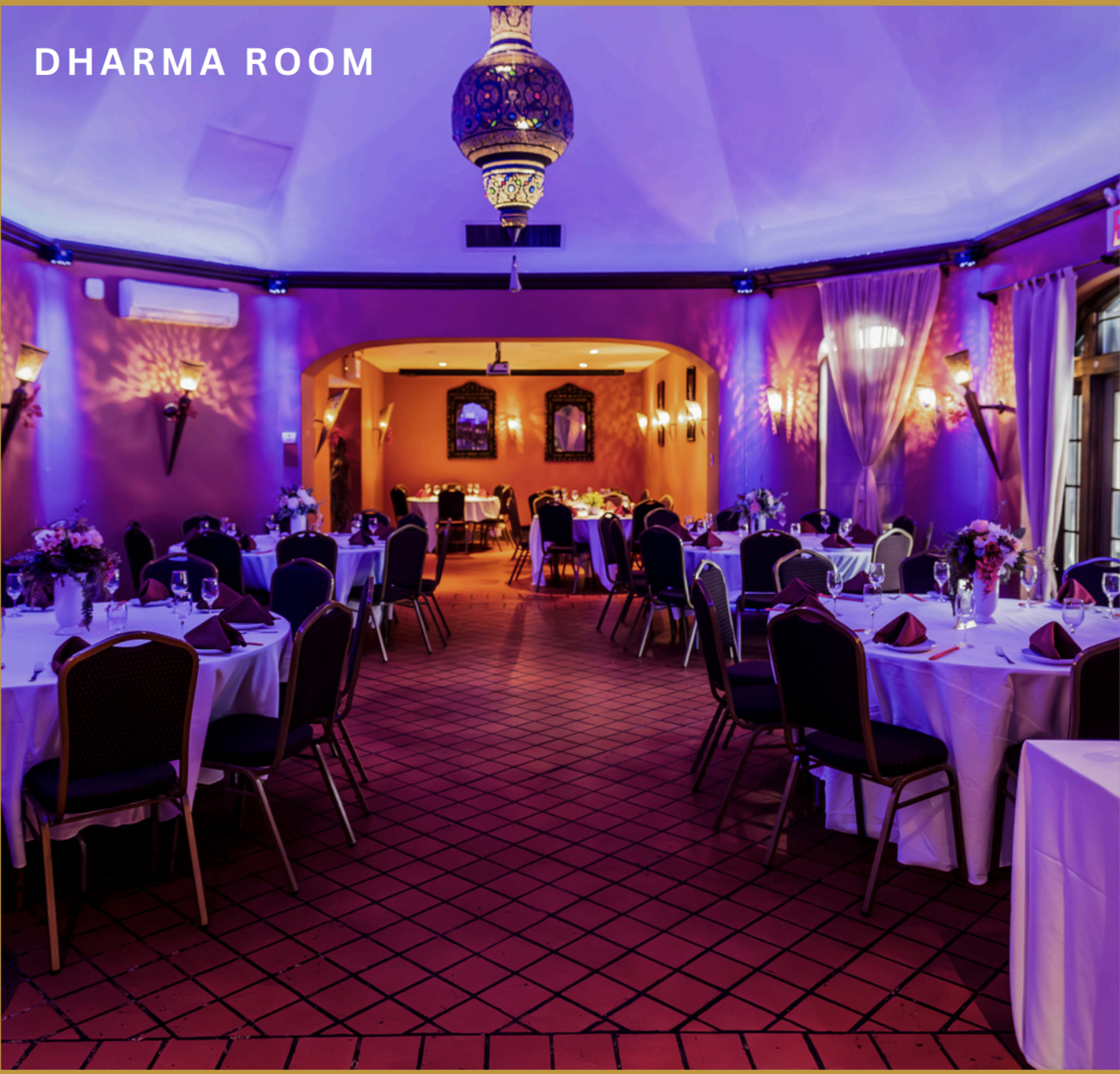 The image shows a dining room set for an event, featuring round tables with white tablecloths and decorative centerpieces, illuminated by soft purple lighting.