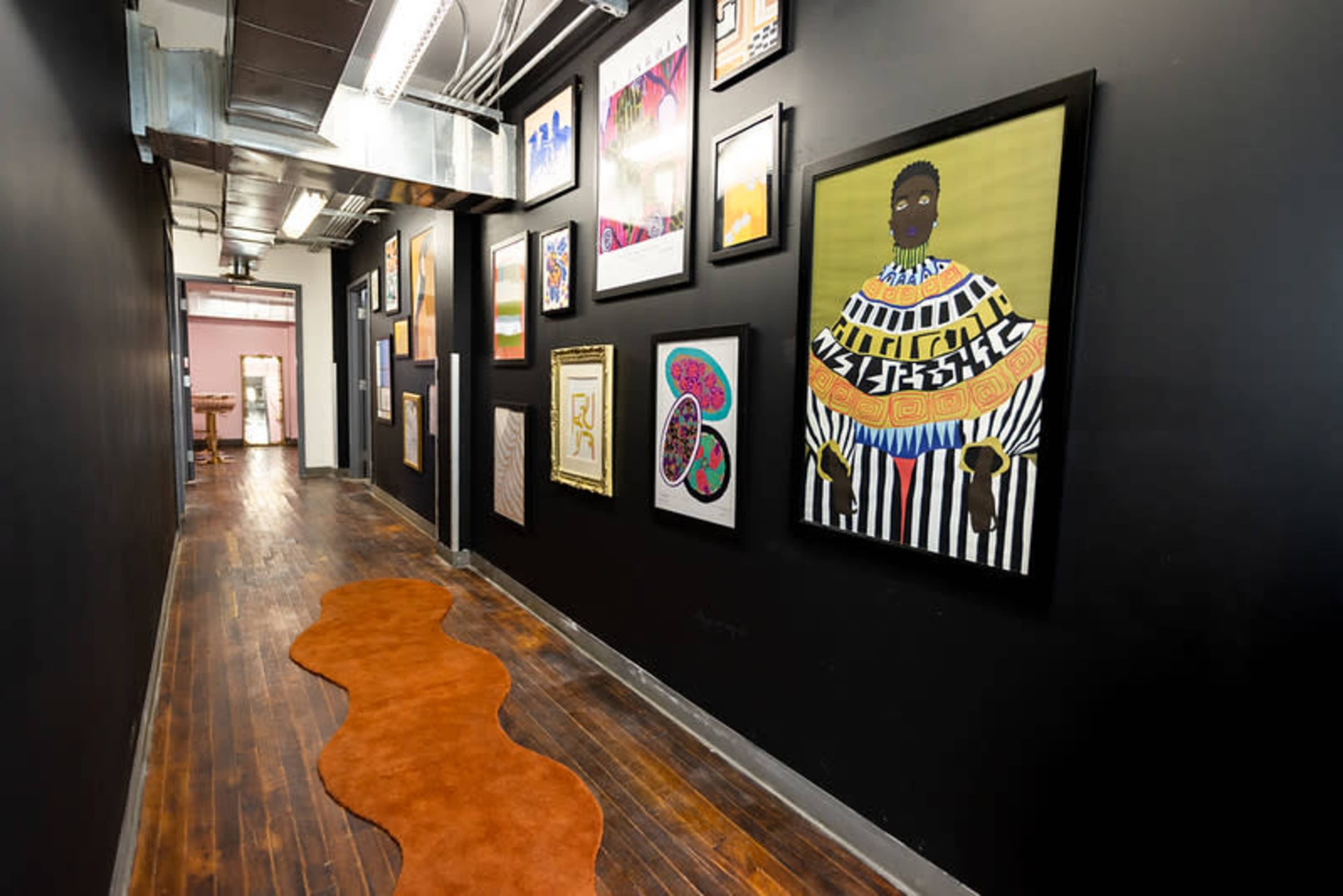 The image shows a narrow hallway lined with framed artworks on black walls, featuring a distinctive orange rug along the wooden floor.