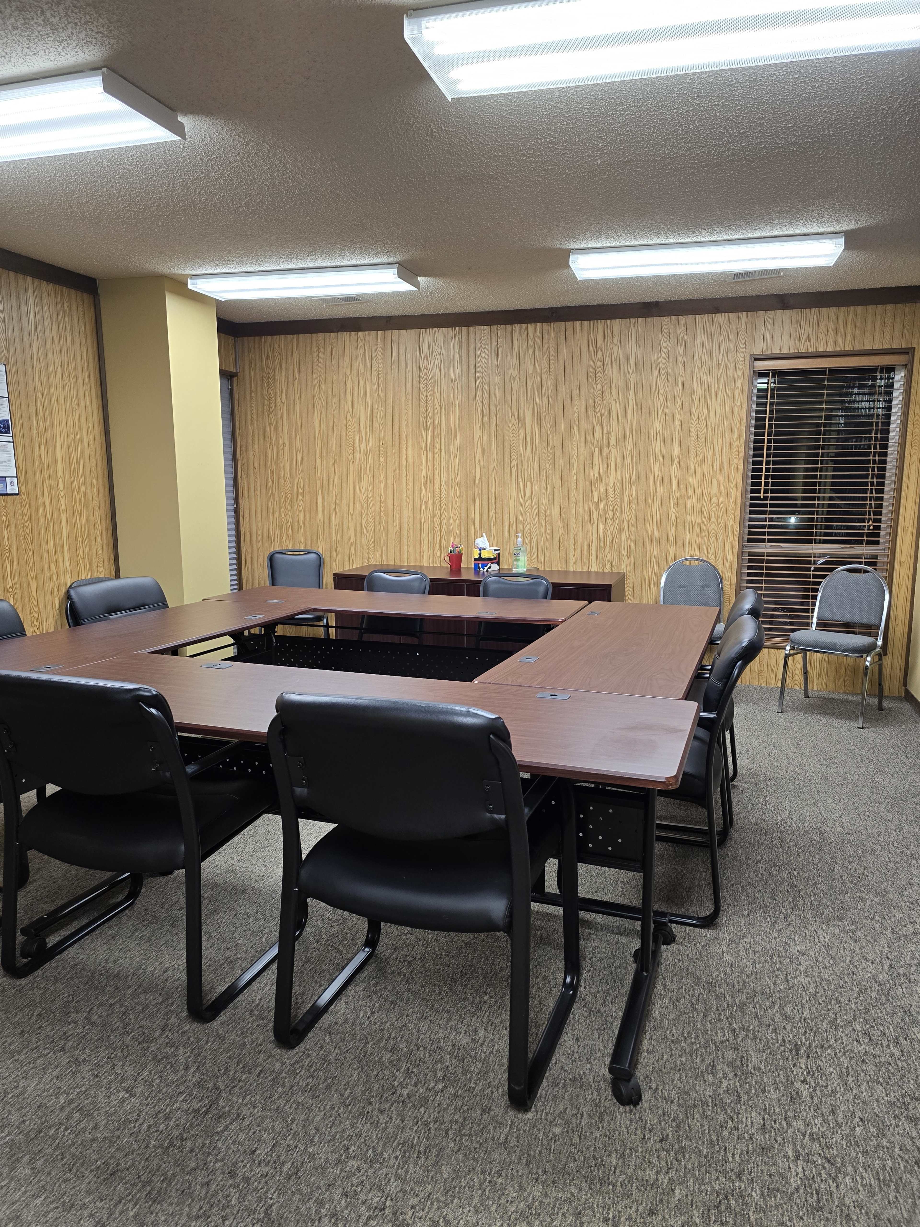 A rectangular conference room features a large table surrounded by black chairs and wooden paneling on the walls.