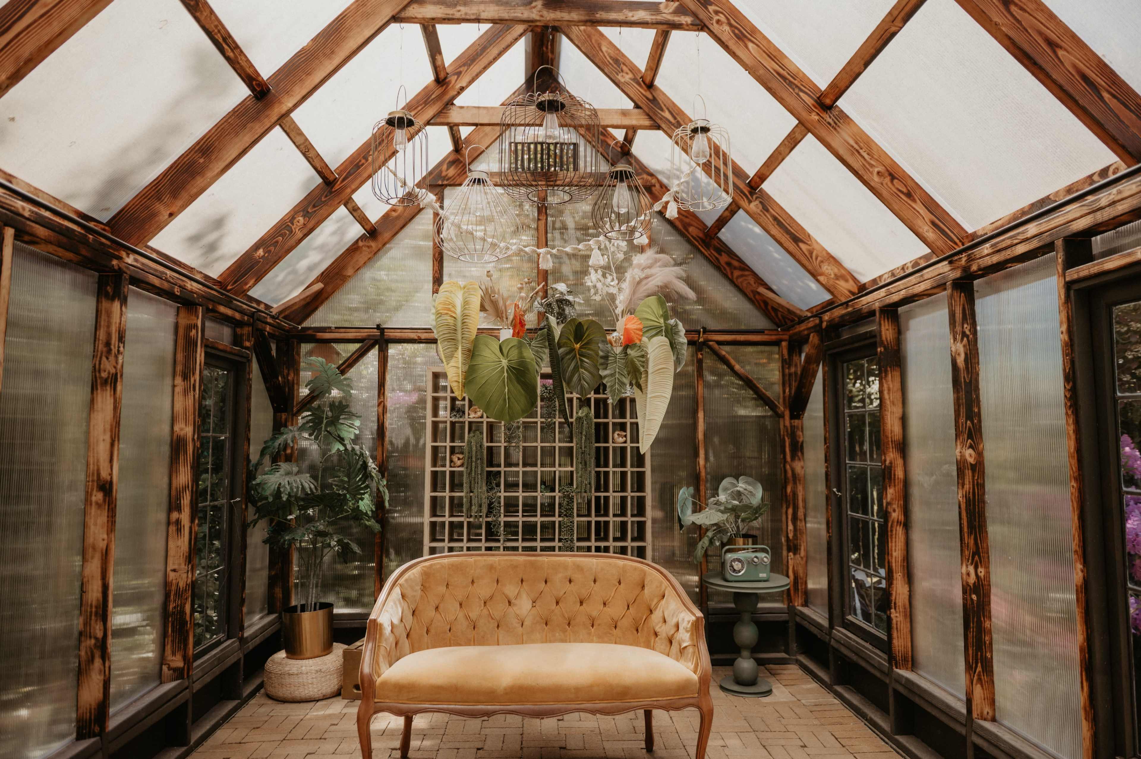 The image shows a greenhouse interior featuring a golden-colored sofa, large green plants, and a decorative chandelier above.