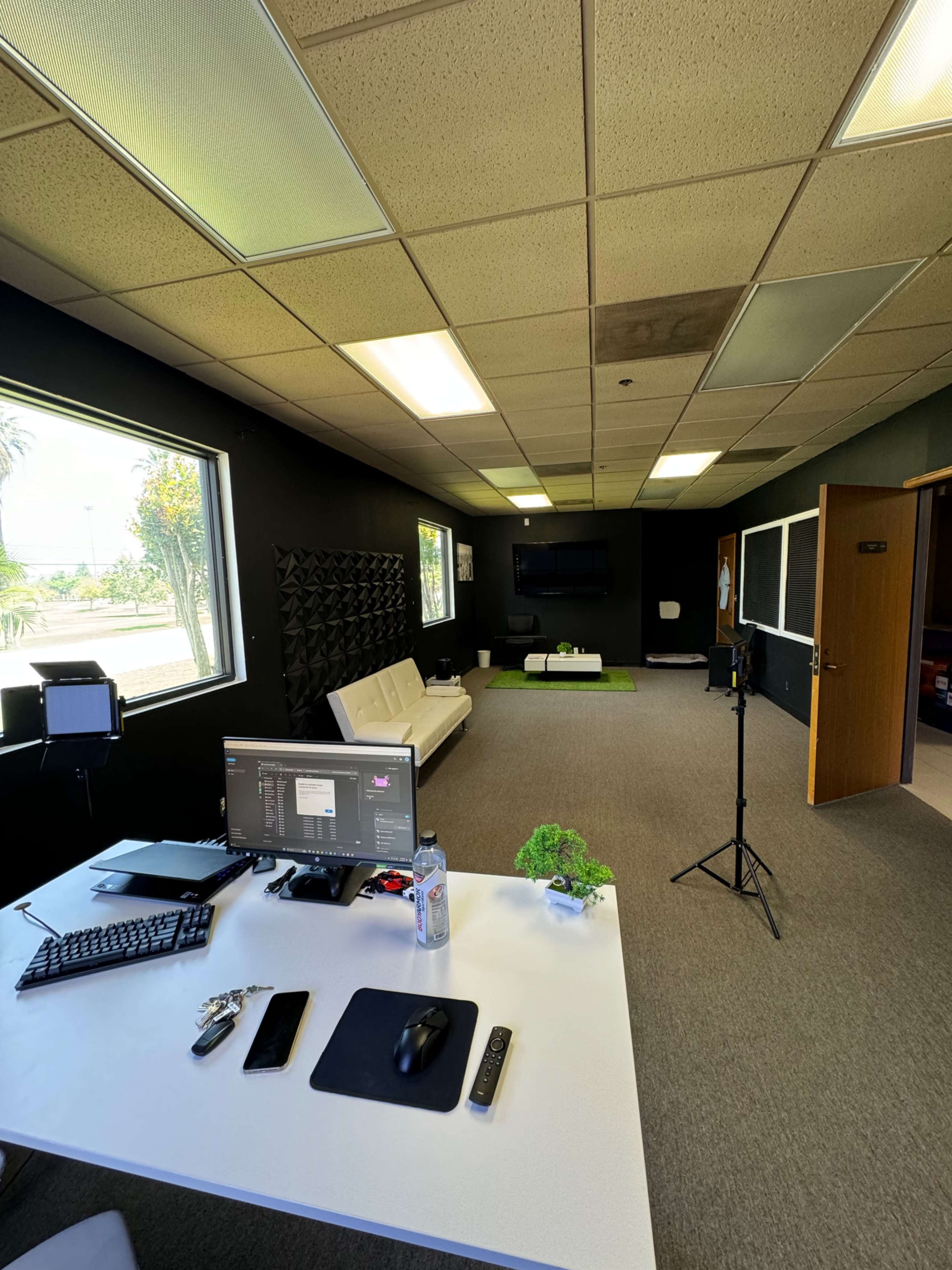 The image shows an office space with a desk featuring a computer, a white couch, and multiple large windows allowing natural light.